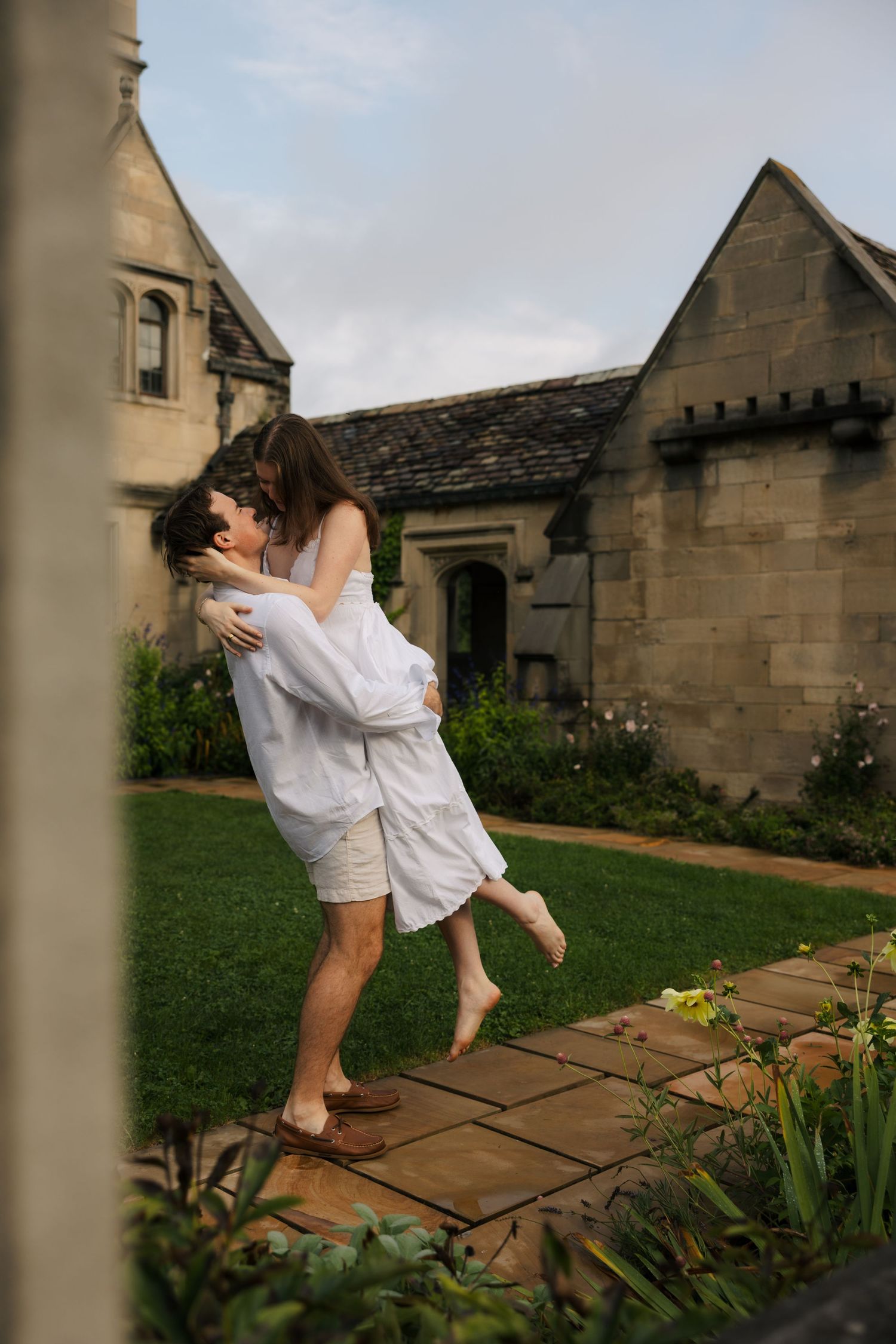 Man lifting woman in white dress in romantic embrace outside stone manor garden.