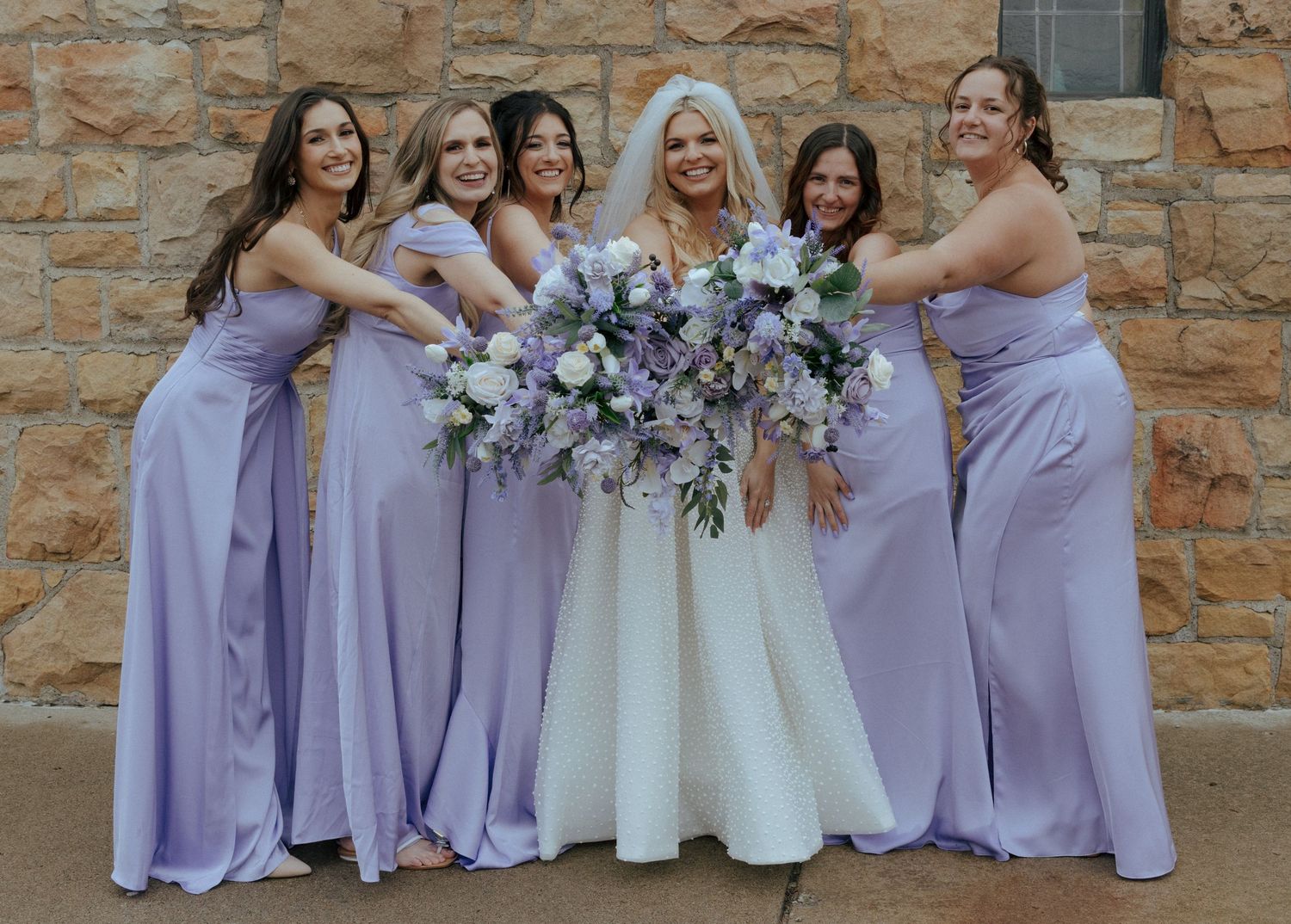 Wedding party in lavender bridesmaid dresses poses together against a stone wall.