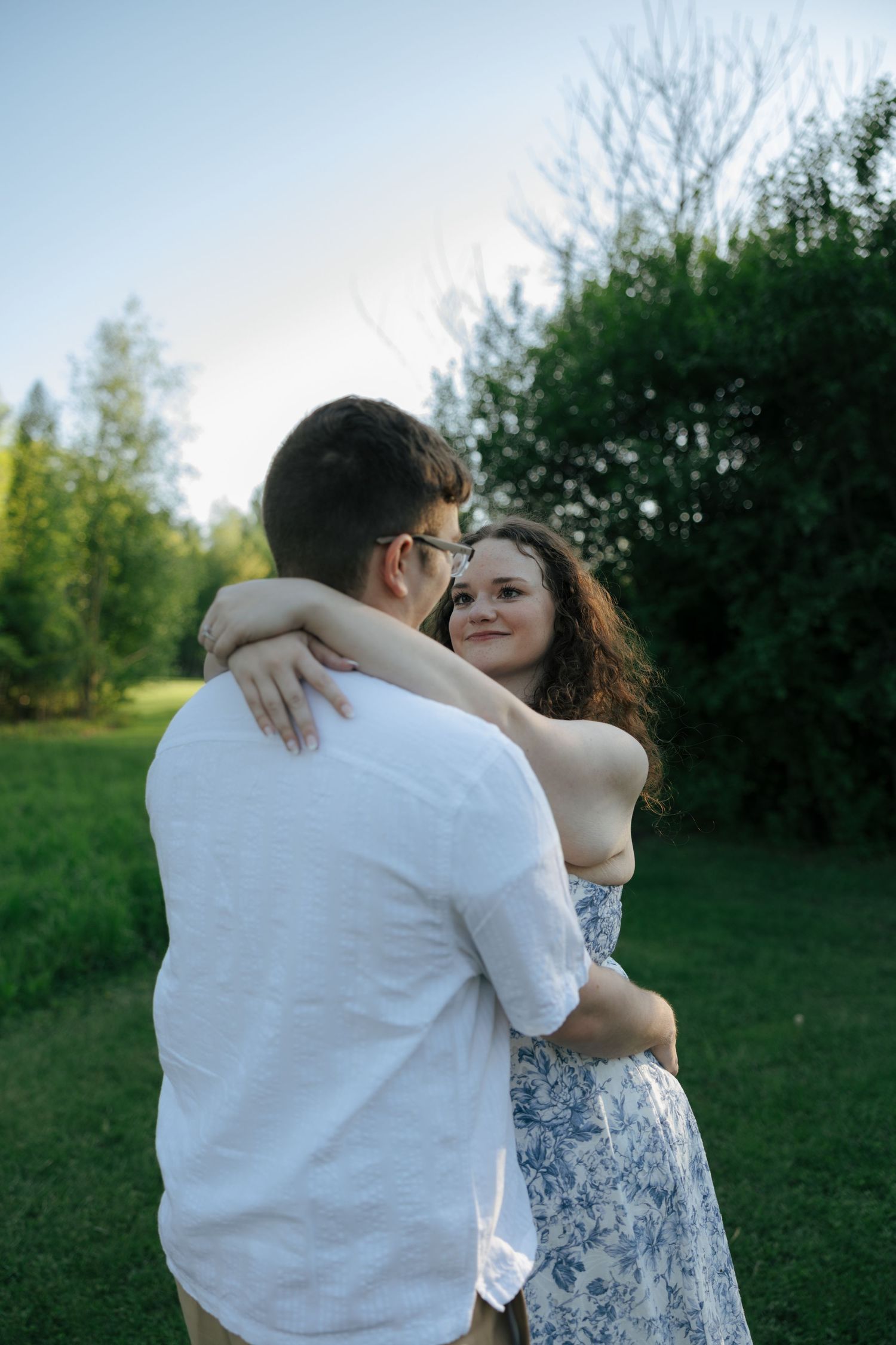 Woman in blue floral dress embracing man in white shirt in a green park, smiling warmly during engagement photos.