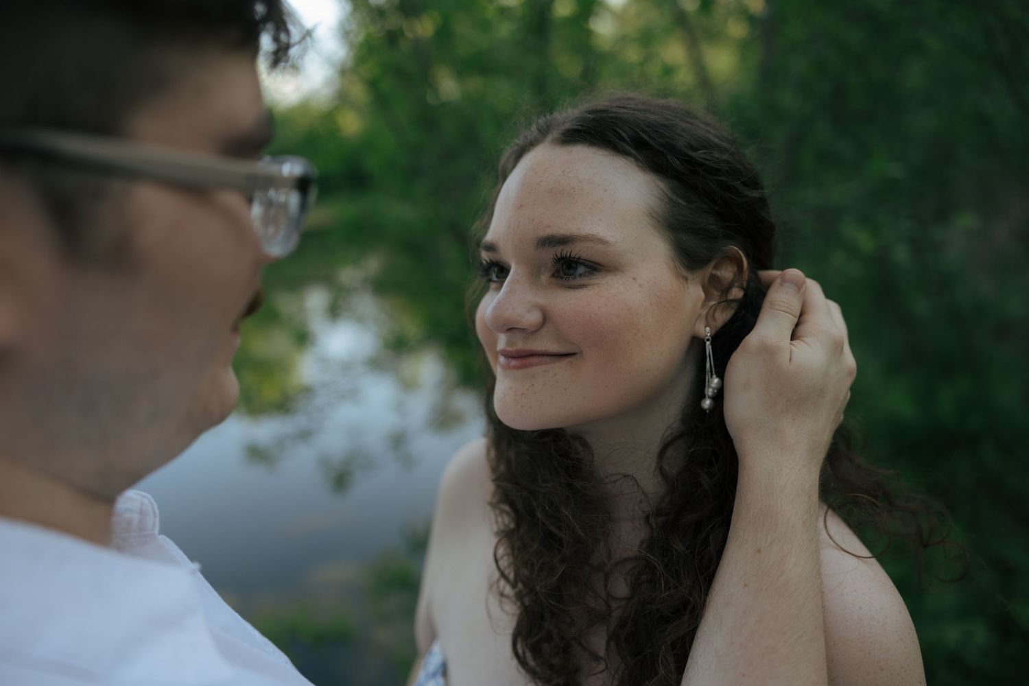 A man gently tucks hair behind a smiling woman's ear outdoors, showcasing elegant drop earrings in a romantic moment.