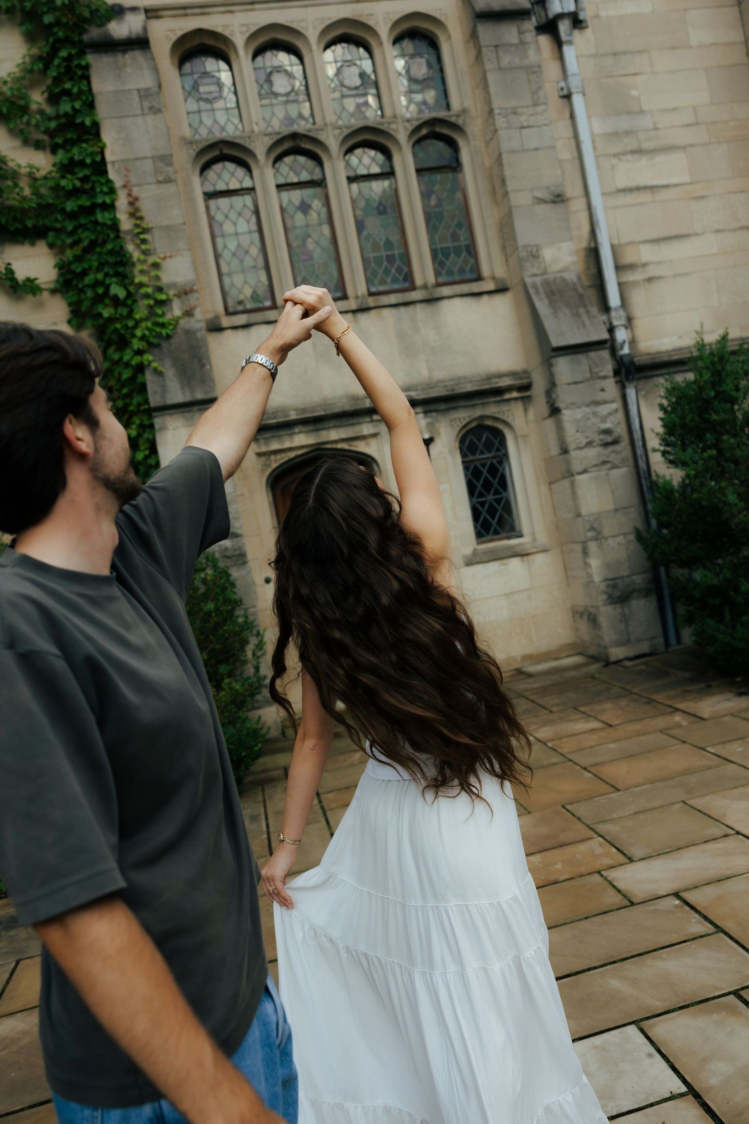 A man twirls a woman in a white dress outside a Gothic stone building with tall windows.