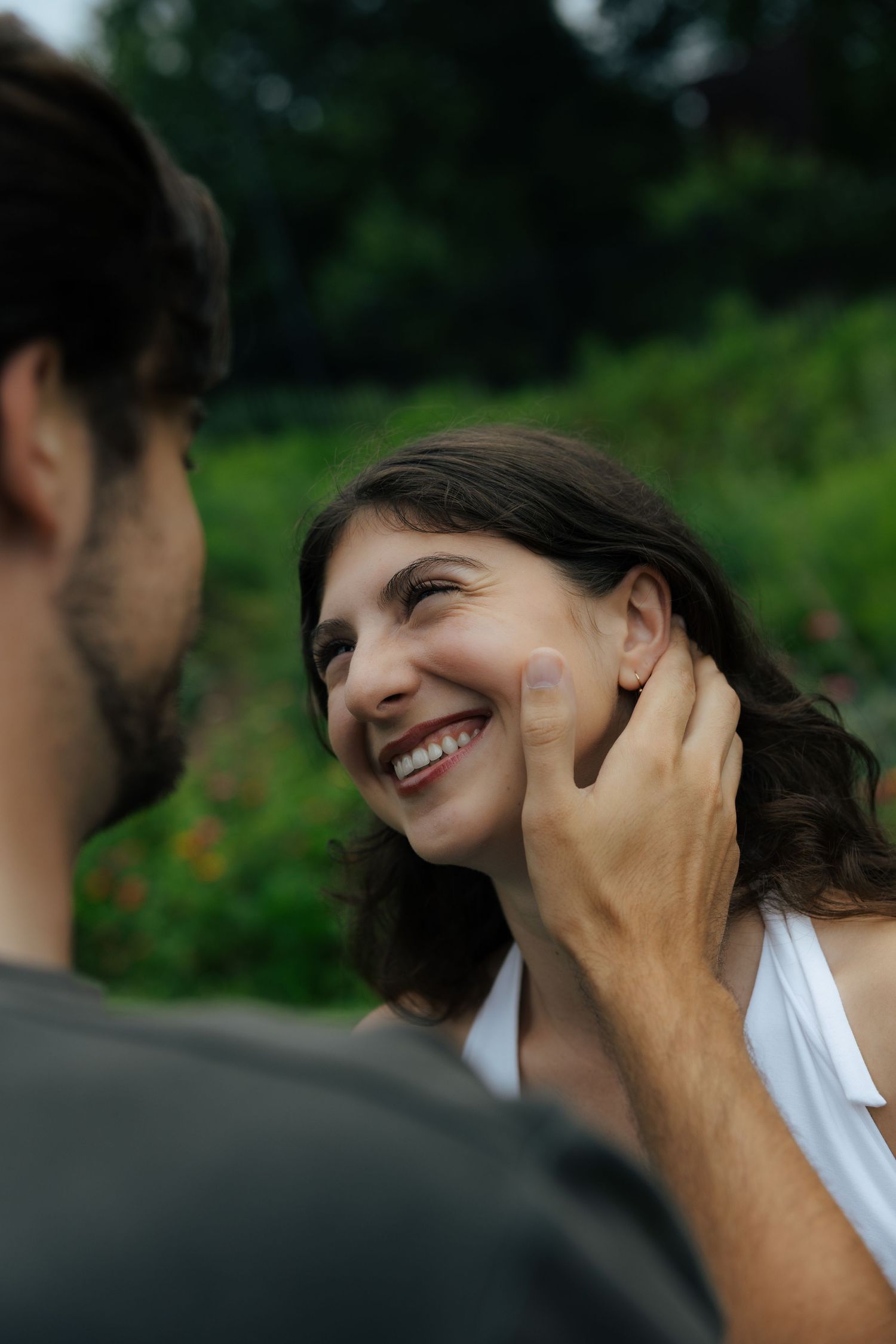 A joyful woman in a white dress laughs as a man gently touches her face outdoors in a lush green garden.