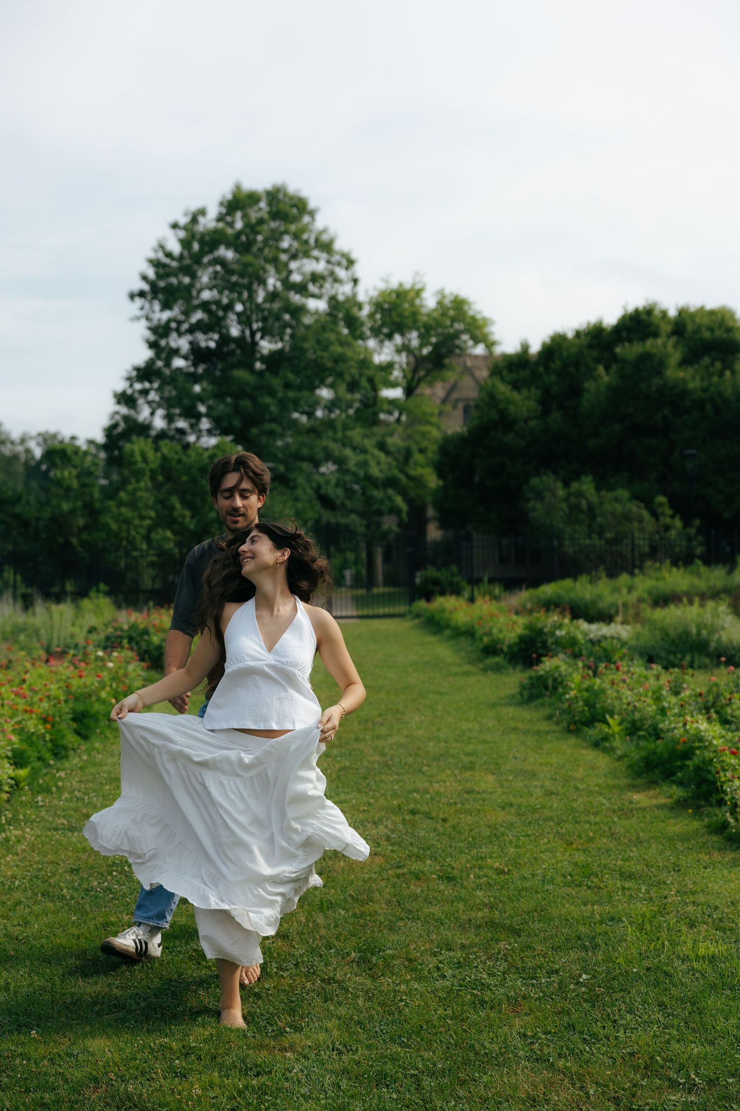 A carefree couple walks together on a grassy garden path, the woman in a flowing white dress laughing.