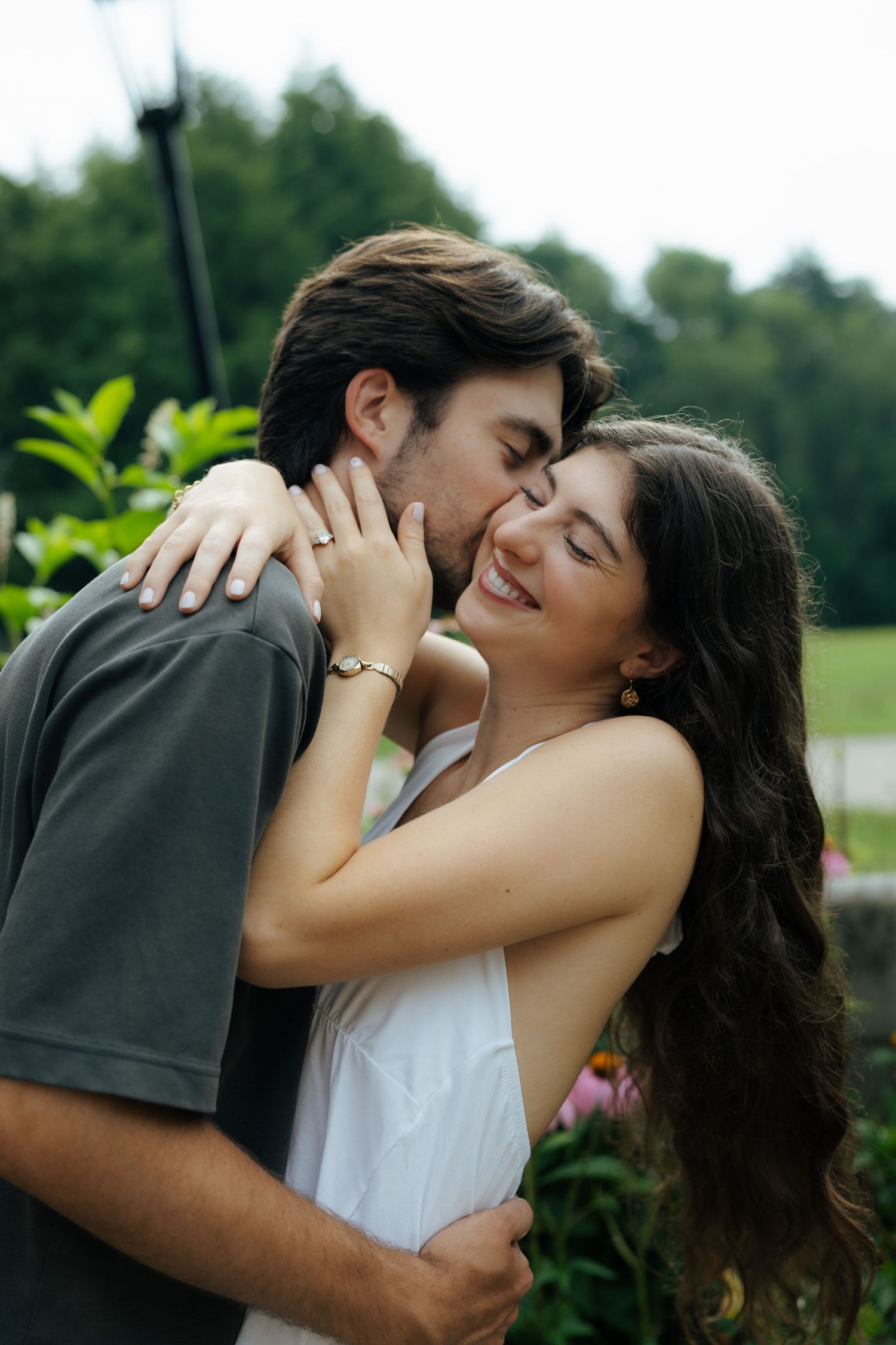Man kissing womans cheek as she laughs happily while embracing outdoors near lush green garden.