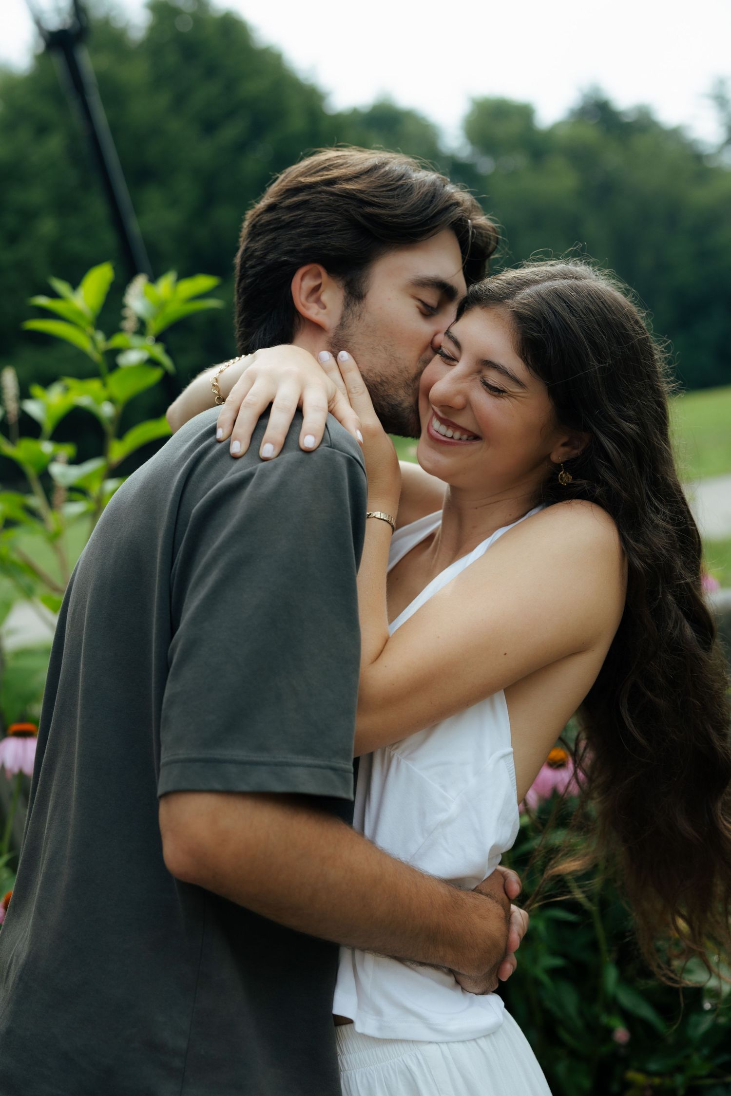 Couple sharing a joyful embrace with man kissing womans cheek near colorful outdoor flower garden.