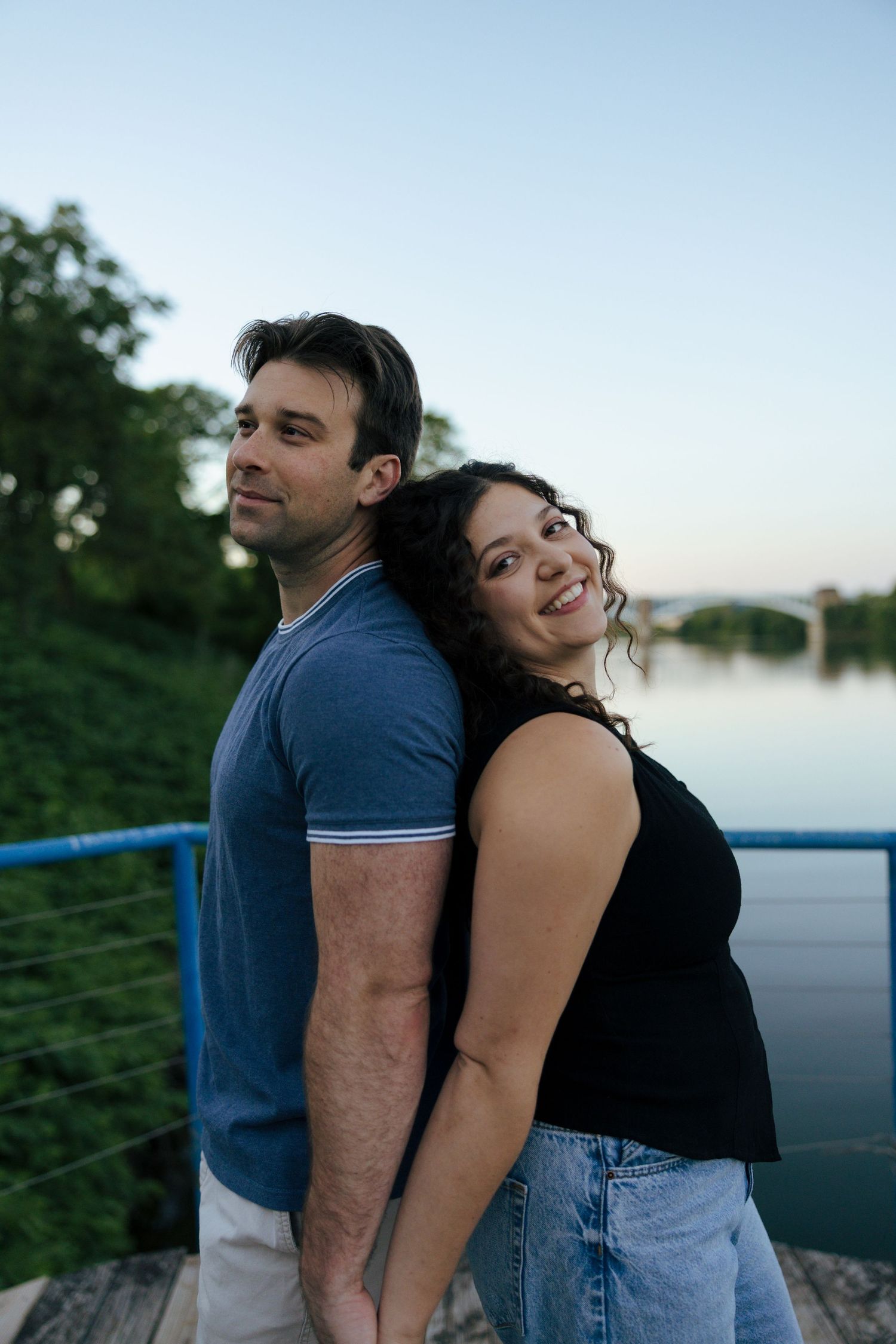 Couple posing back-to-back by a river at dusk, woman smiling at camera in black top and jeans.