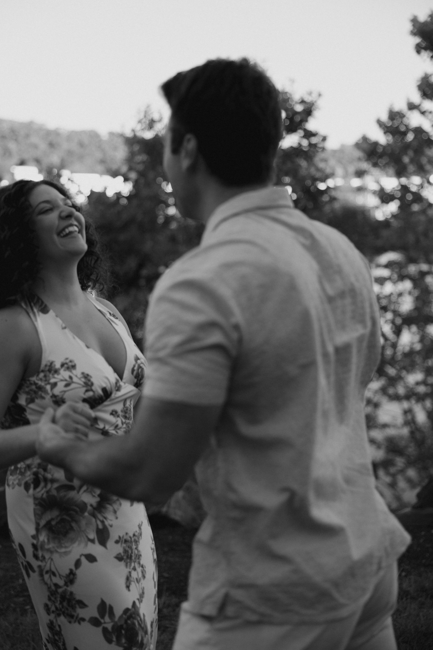 A black and white photo of a laughing woman in a floral dress holding hands with a man outdoors.