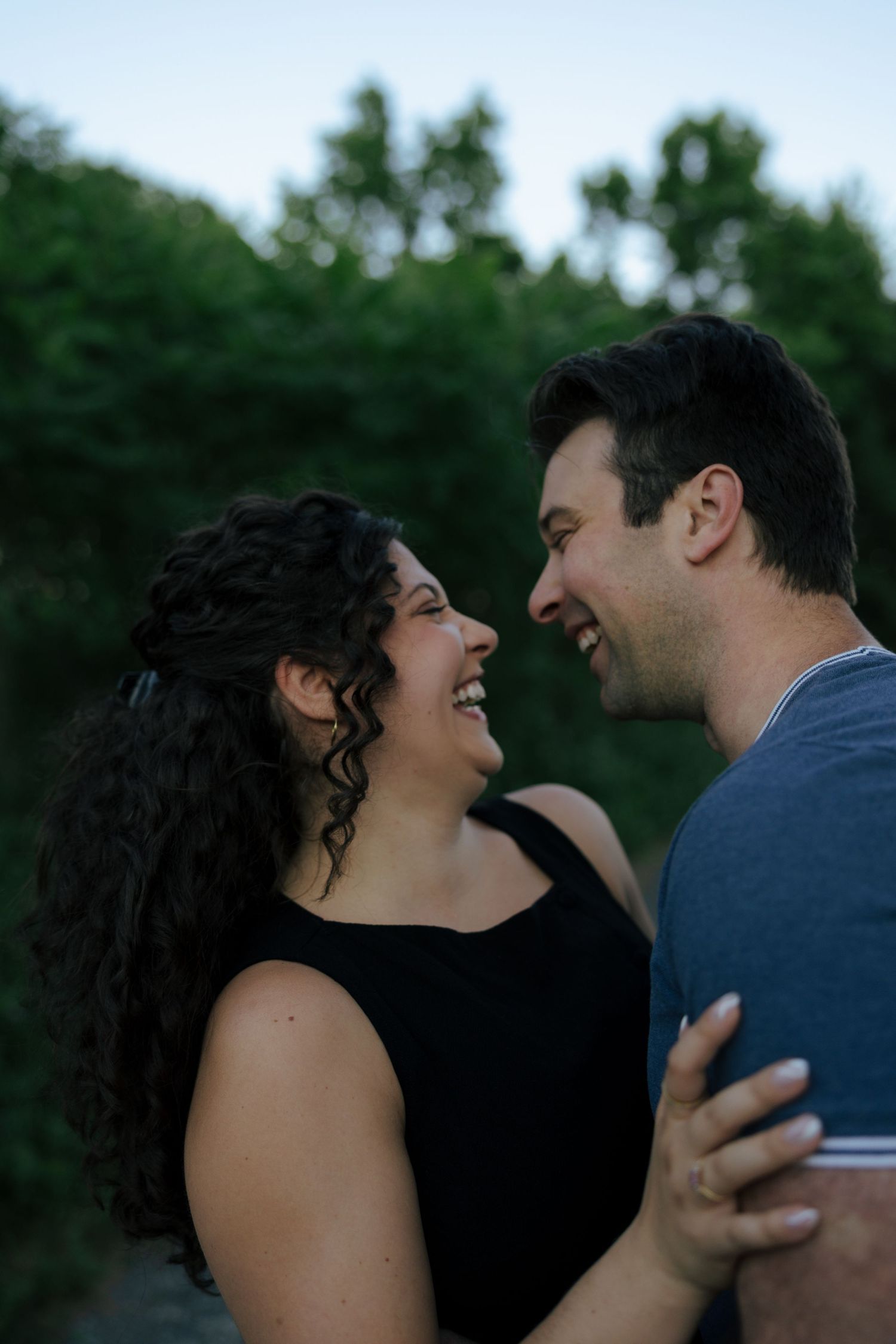 A laughing couple embraces outdoors at dusk, sharing a joyful moment with lush green trees in the background.