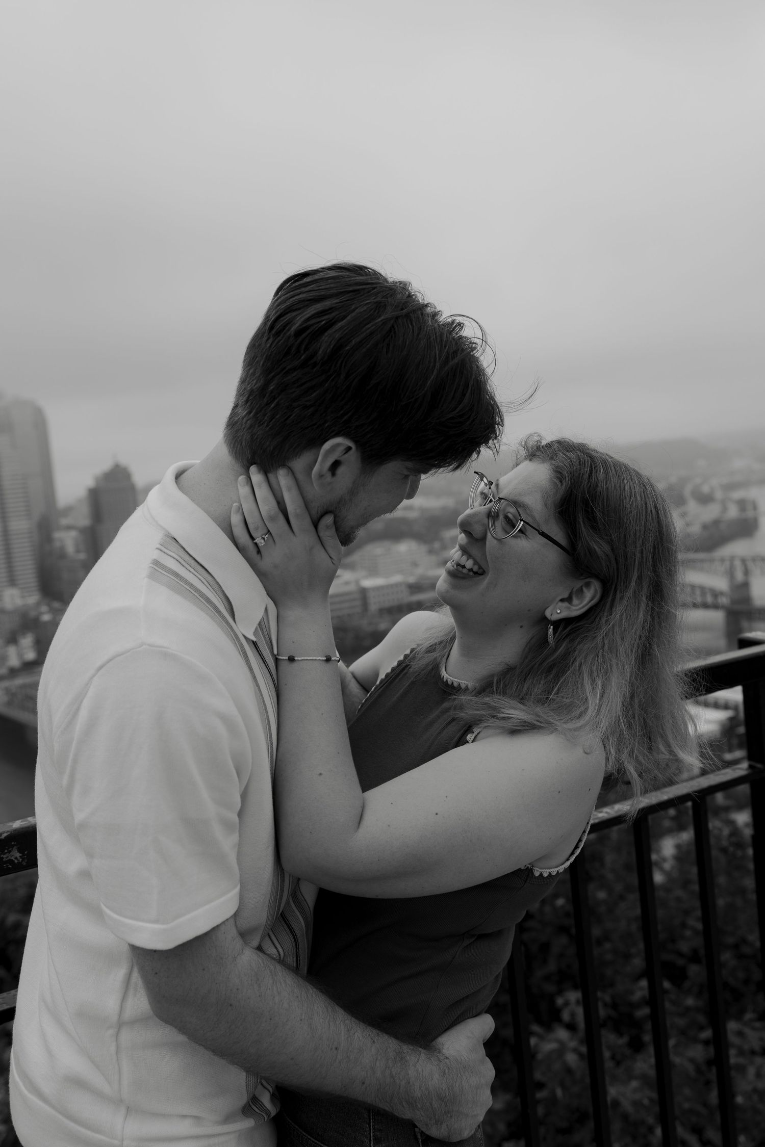 Black and white photo of a smiling couple sharing a tender moment with a city skyline in the background.