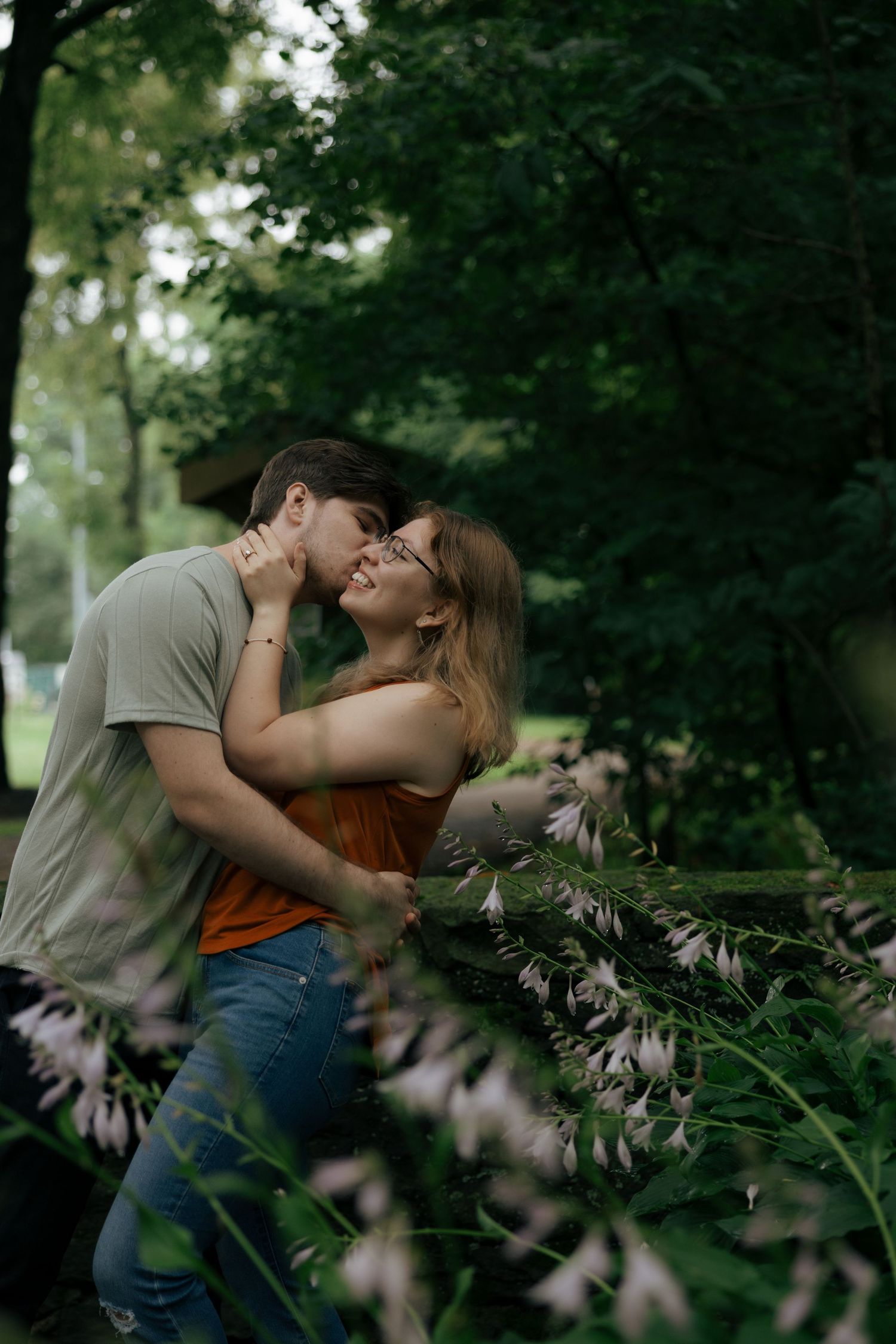 A couple shares a romantic kiss surrounded by lush green trees and blooming flowers in a serene park setting.