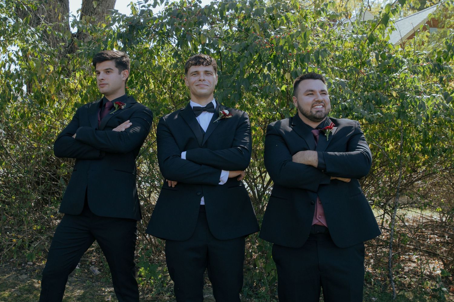 Three groomsmen in black suits pose with arms crossed outdoors among green trees.