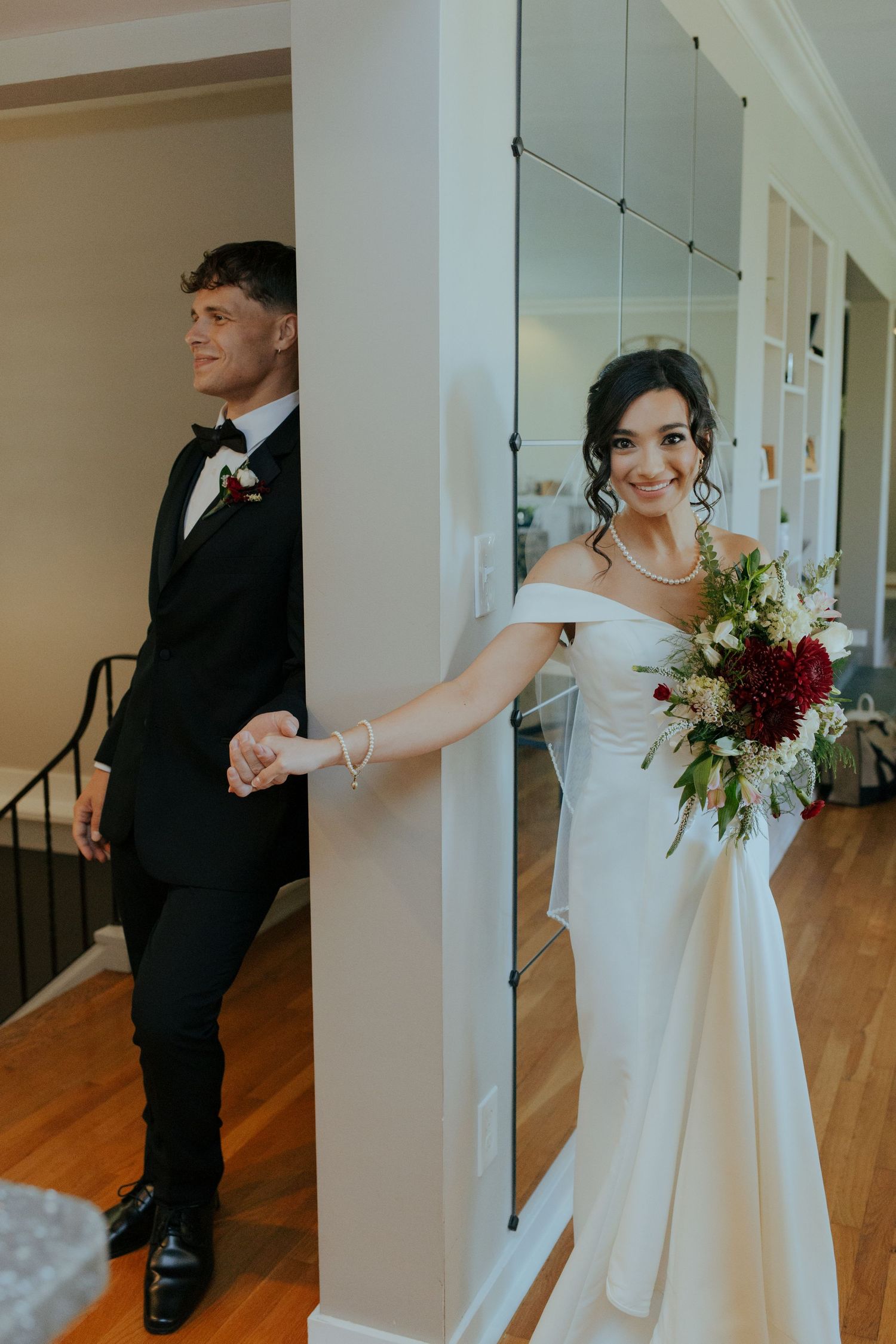Bride and groom share a tender first look moment, holding hands around a white column, bride holding a red and white bouquet.