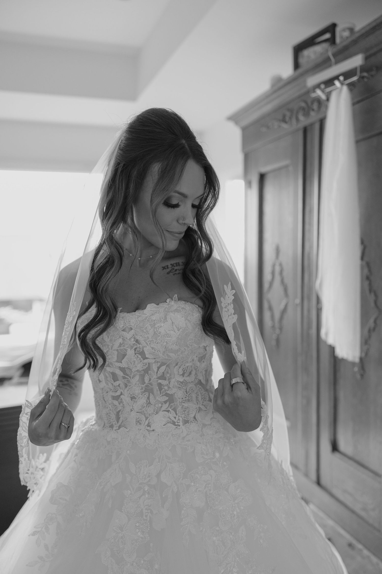 A bride in a floral lace wedding gown and veil poses elegantly in a black and white portrait.