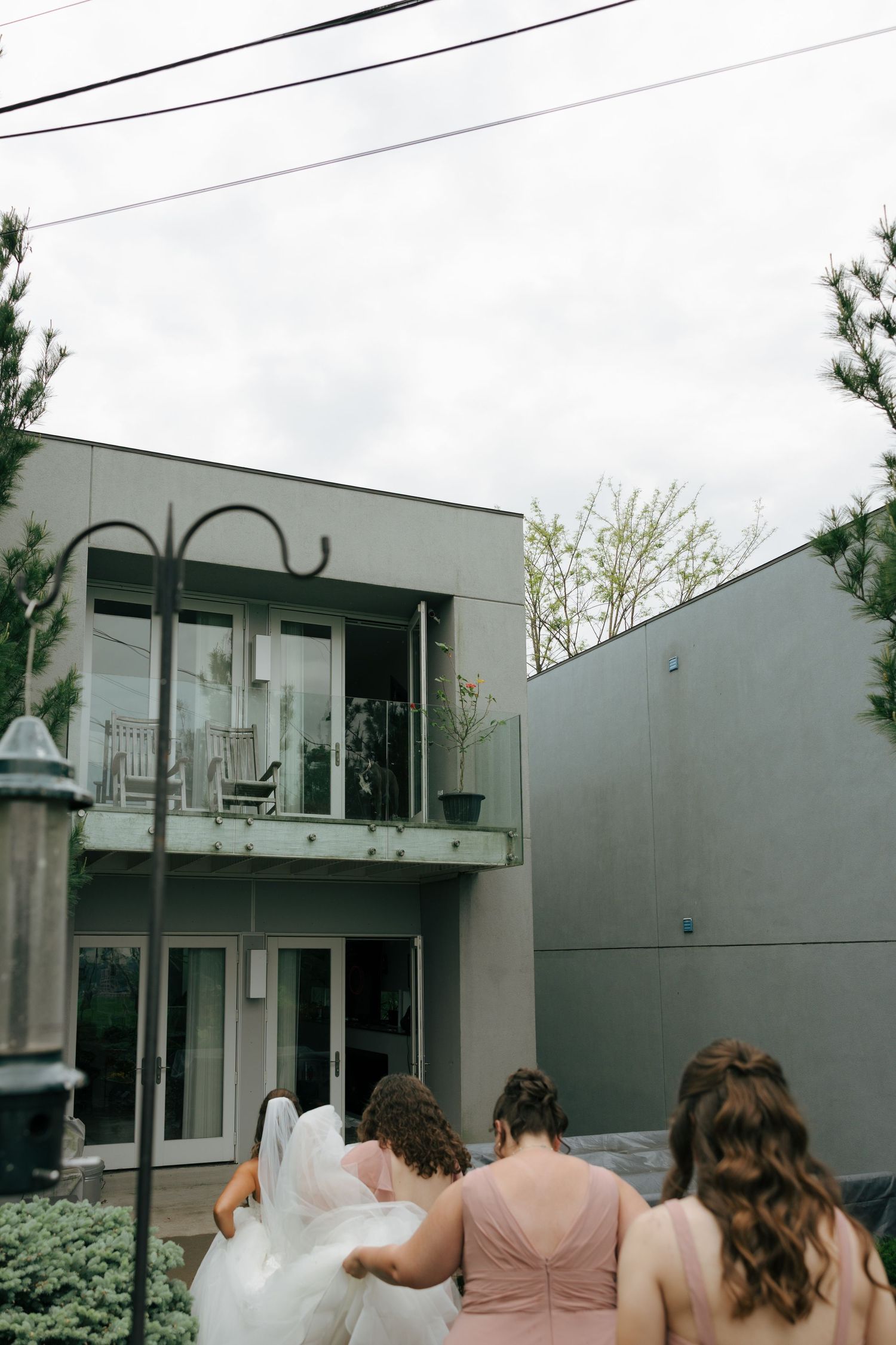 Bridesmaids helping bride with veil outside modern gray building on cloudy wedding day.