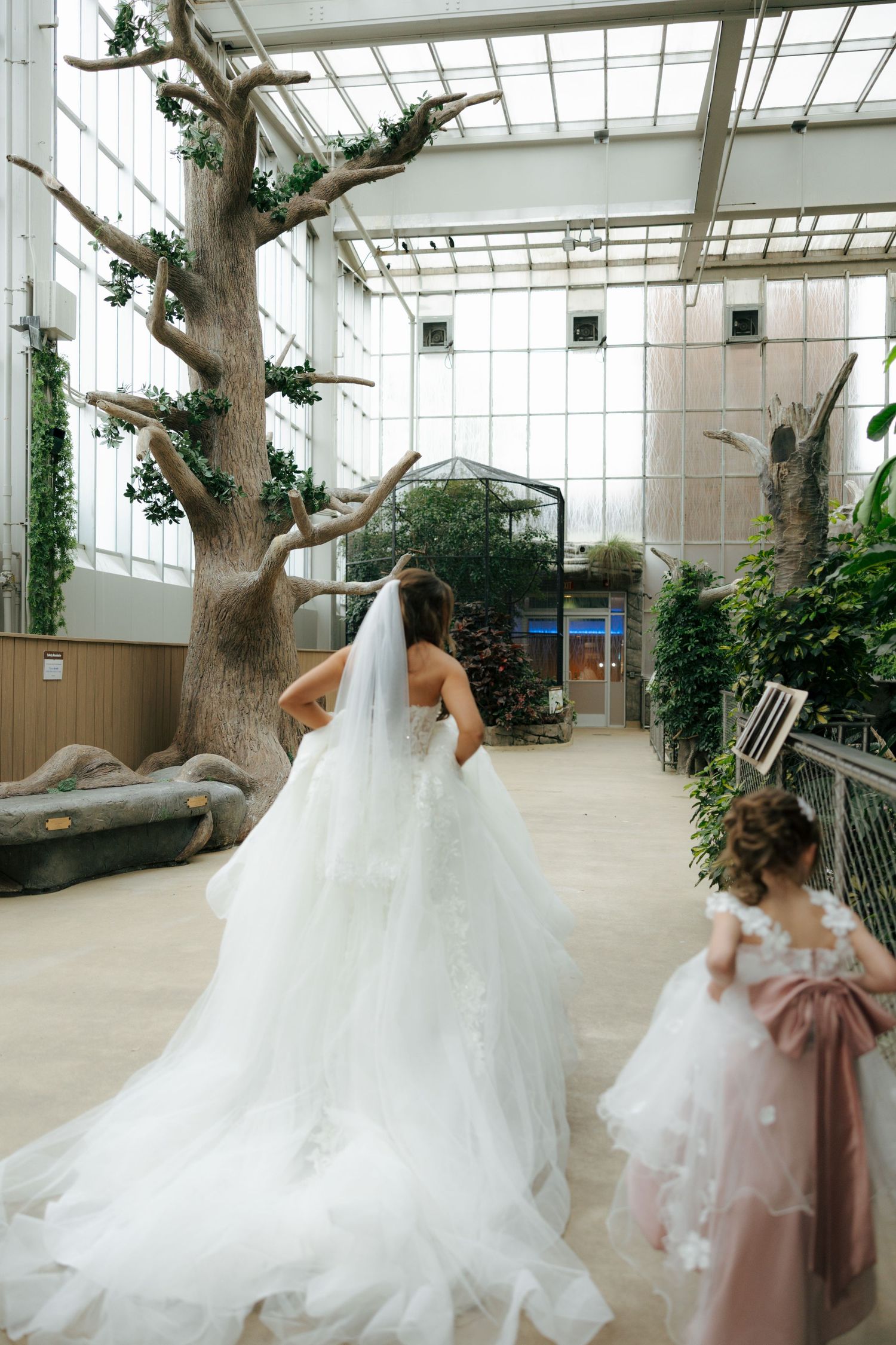 Bride in white gown and veil walks with flower girl in pink dress through an elegant indoor garden venue.