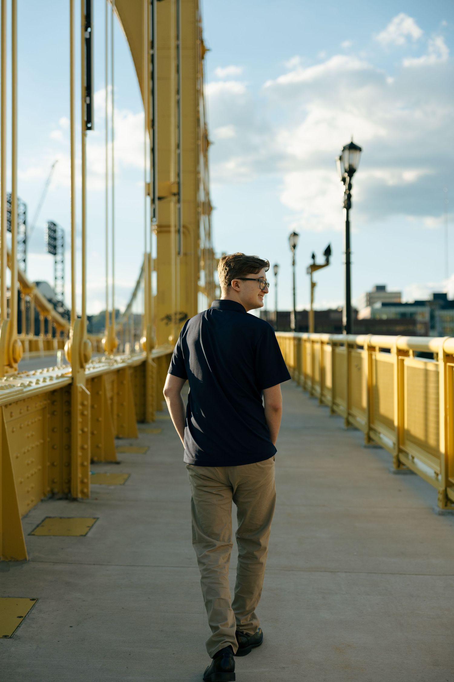 Man in navy polo and khakis walking on yellow suspension bridge pedestrian path on a sunny day.