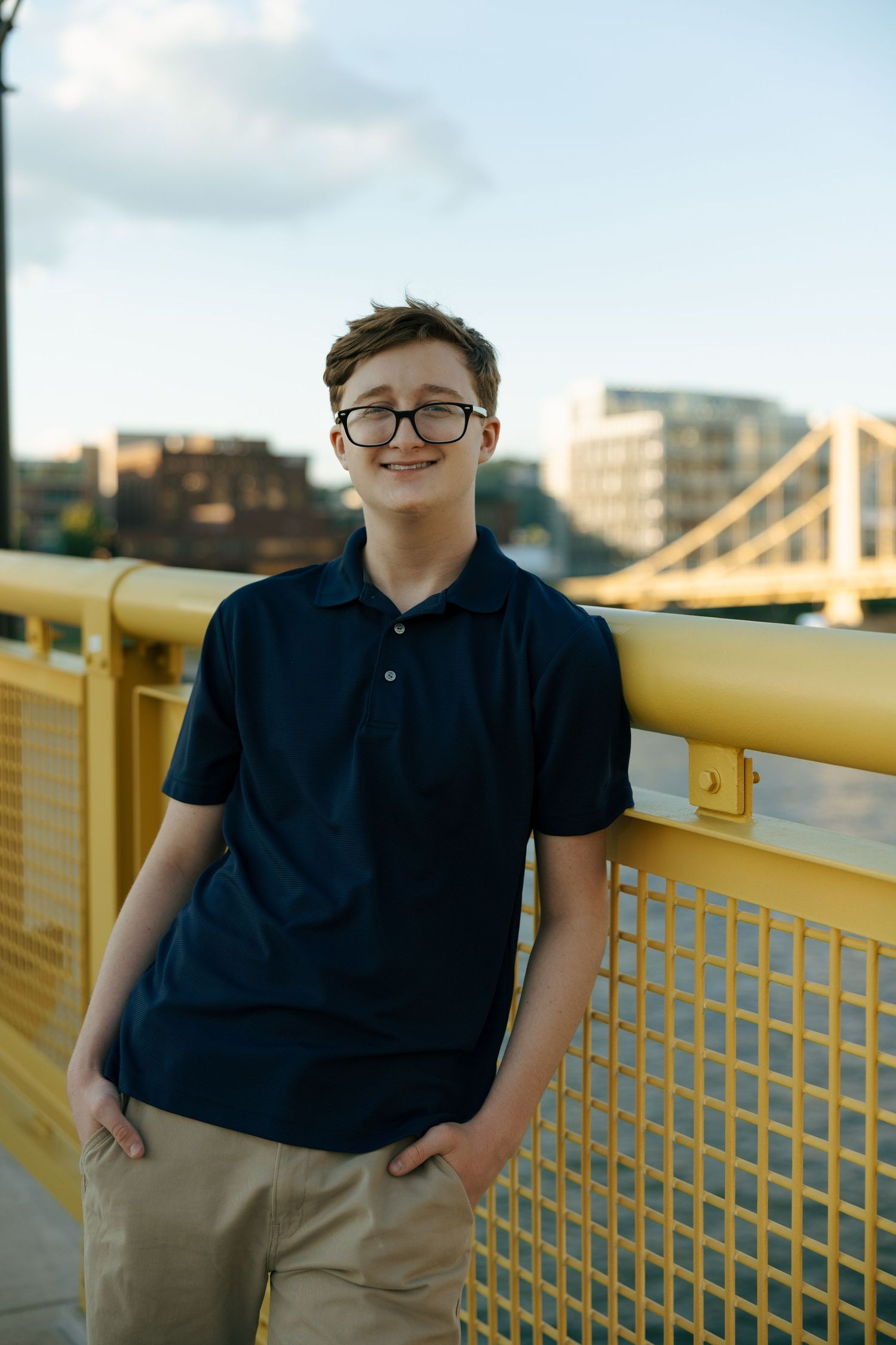 Smiling teen boy with glasses leans on yellow bridge railing in navy polo shirt and khaki pants.