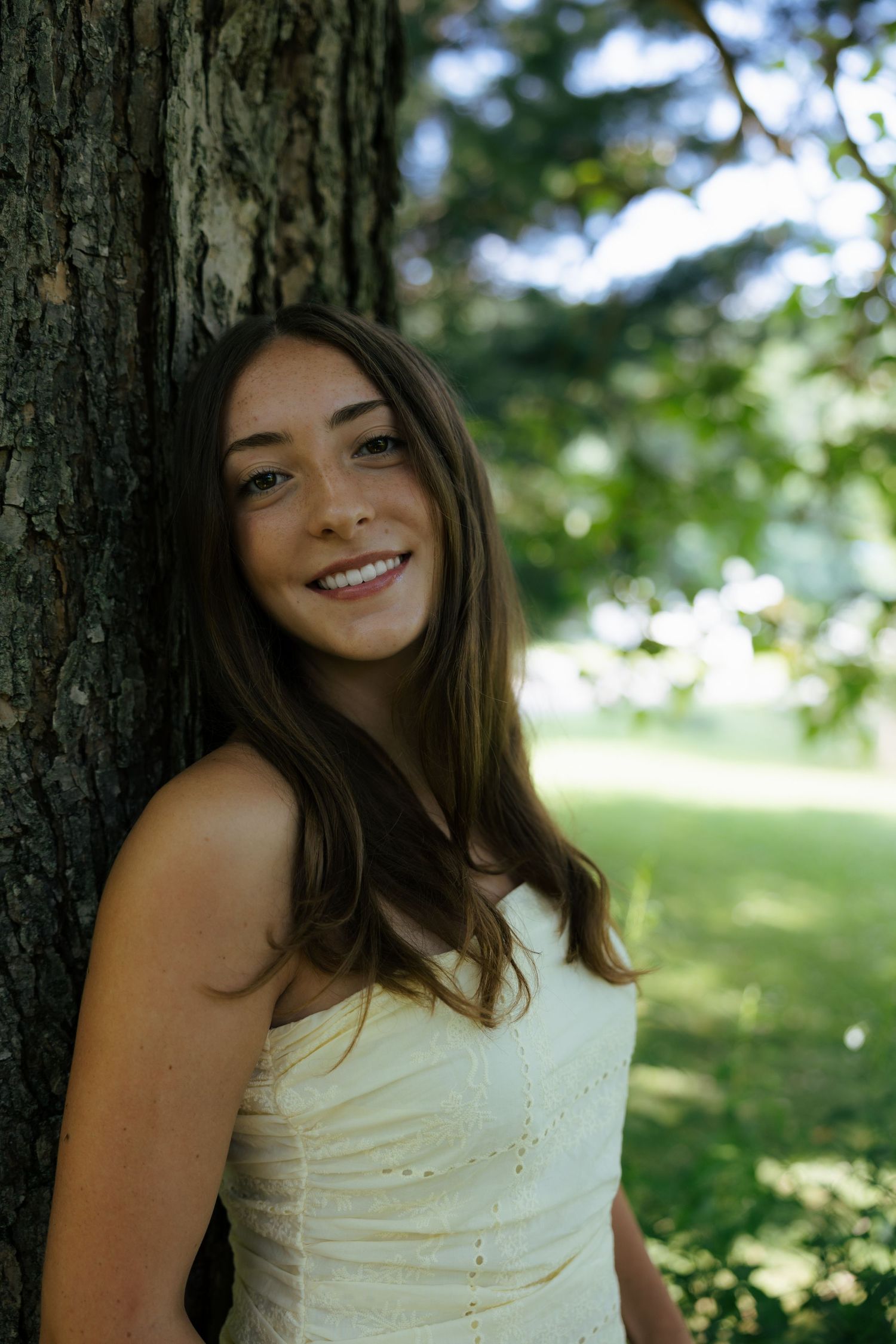 A smiling young woman in a white dress leans against a large tree trunk in a sunny park.