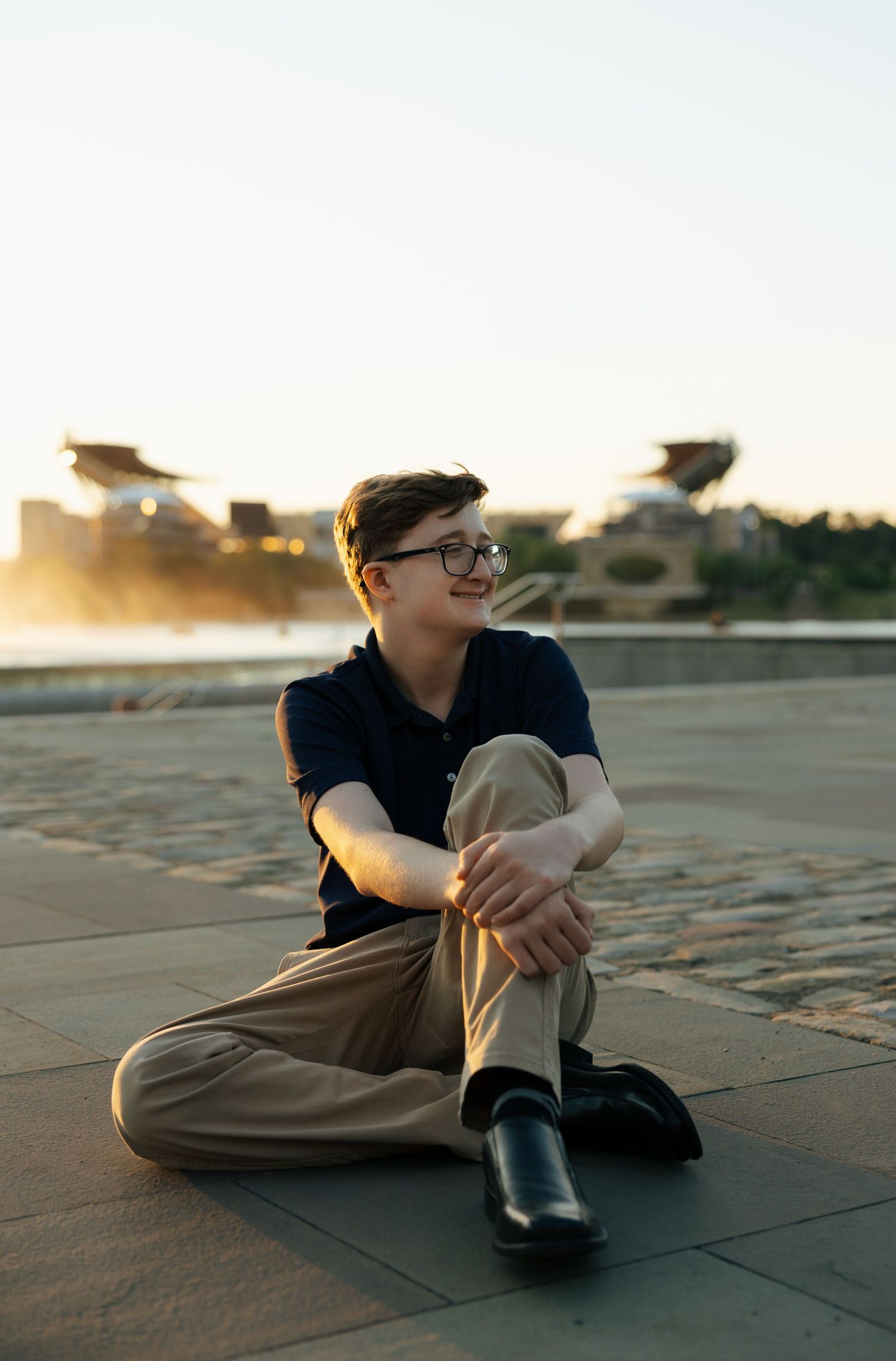Teen boy in navy polo sits cross-legged on plaza tiles at golden hour near waterfront.
