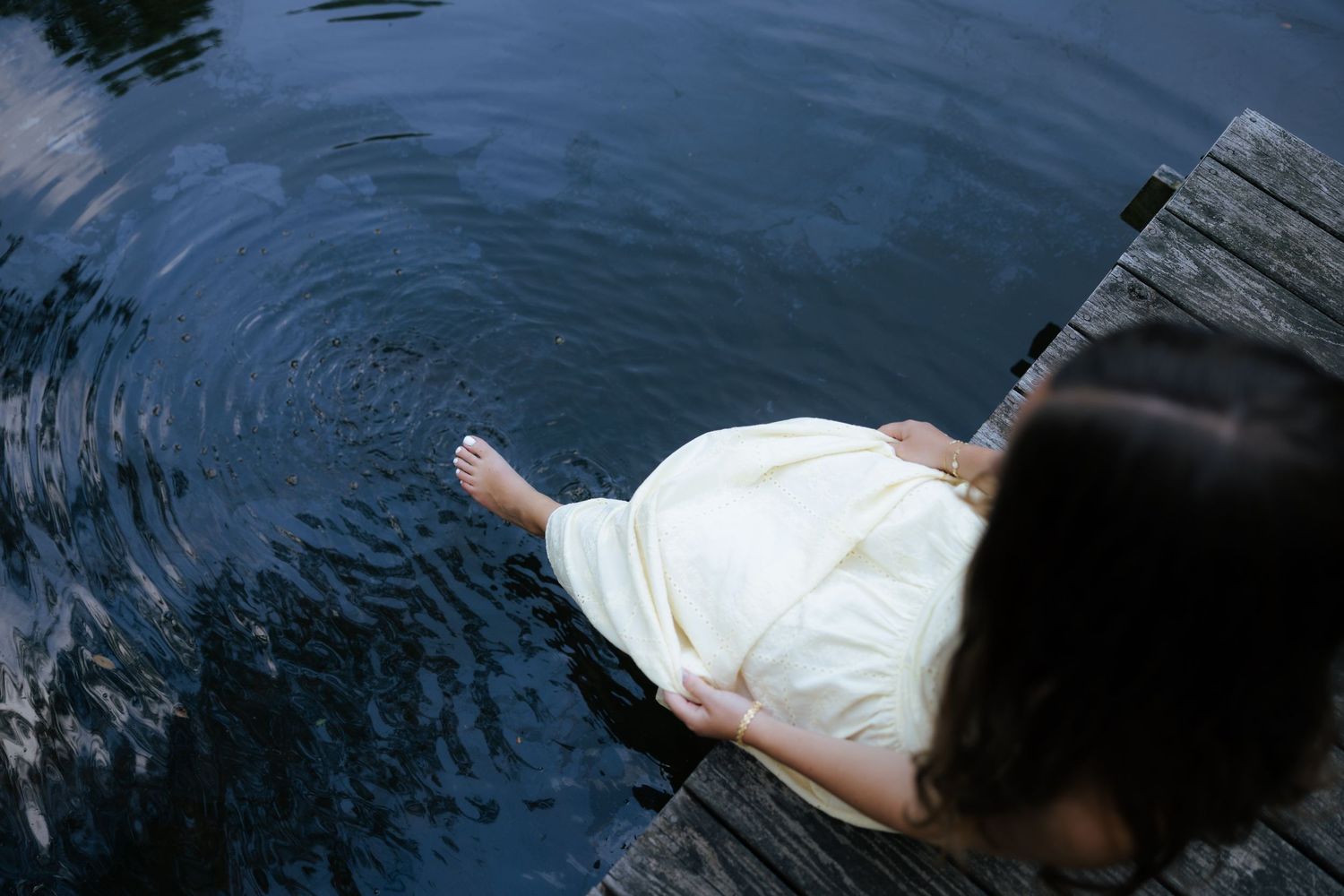Woman in cream dress sitting on dock with feet in calm water.