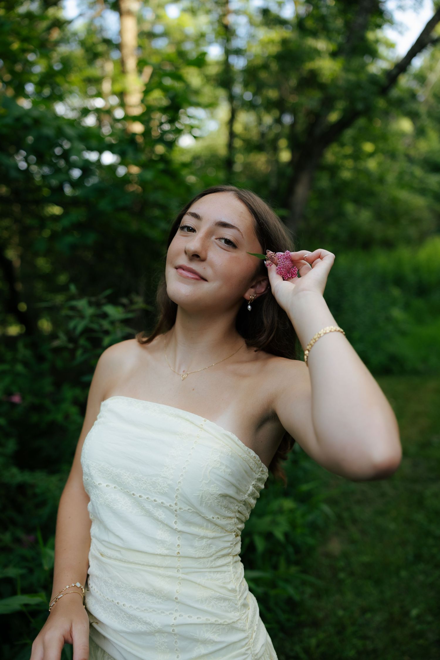 Young woman in strapless white dress holding a pink flower near her ear, smiling in a green garden.