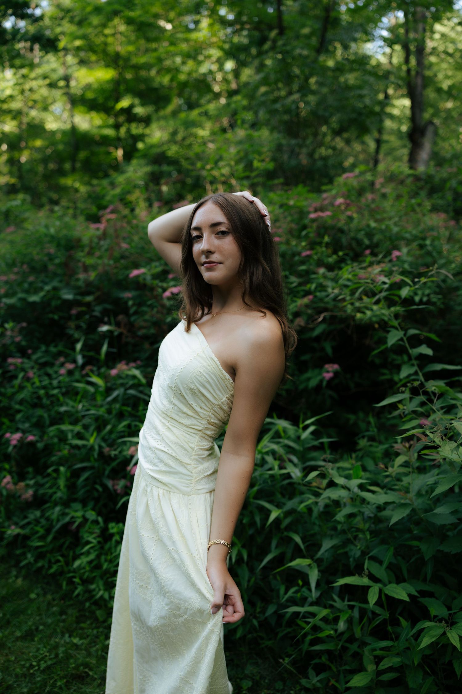 Young woman in cream strapless gown posing among lush green foliage.