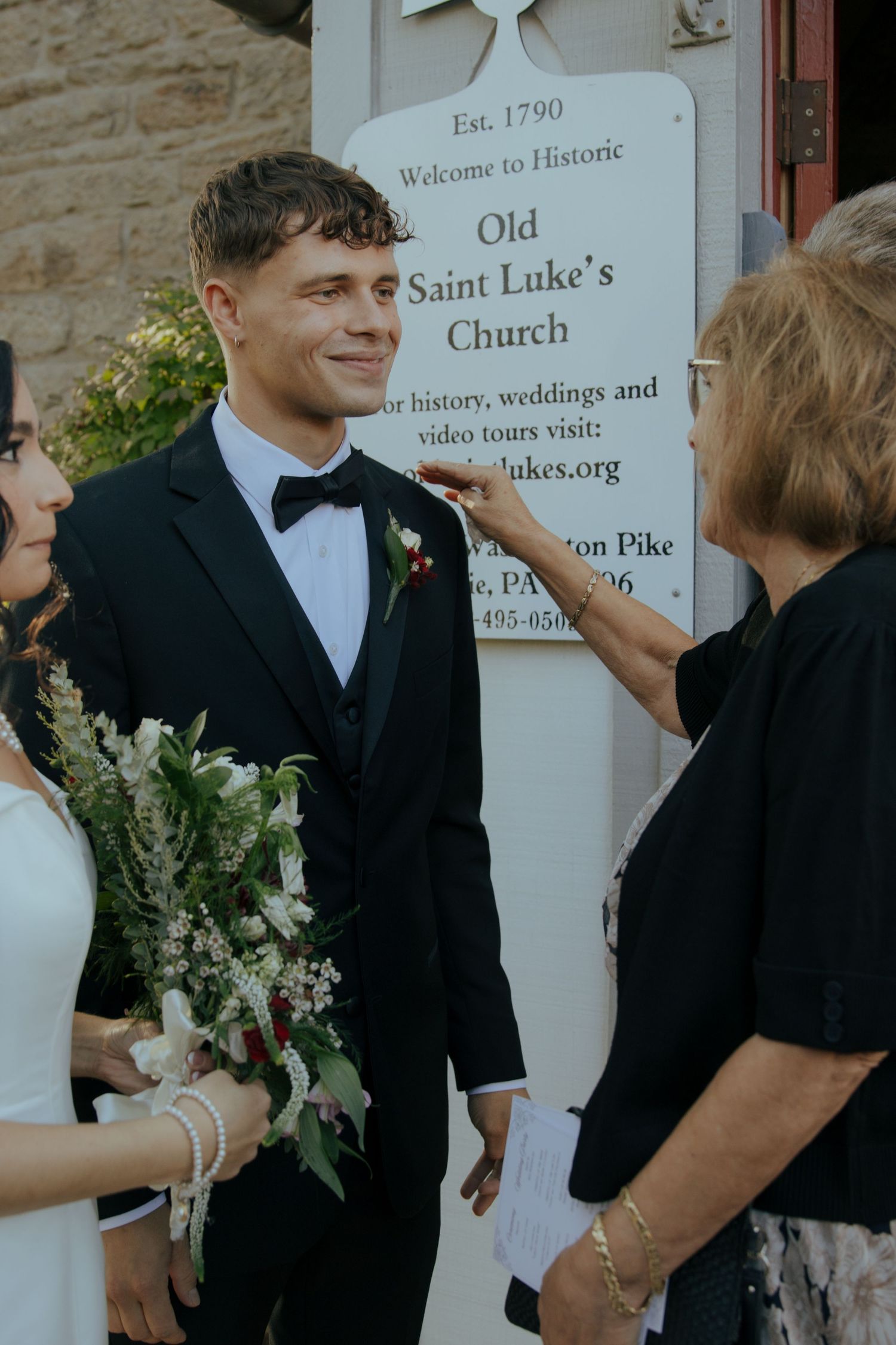 Smiling groom in tuxedo stands with bride outside Historic Old Saint Luke's Church established in 1790.