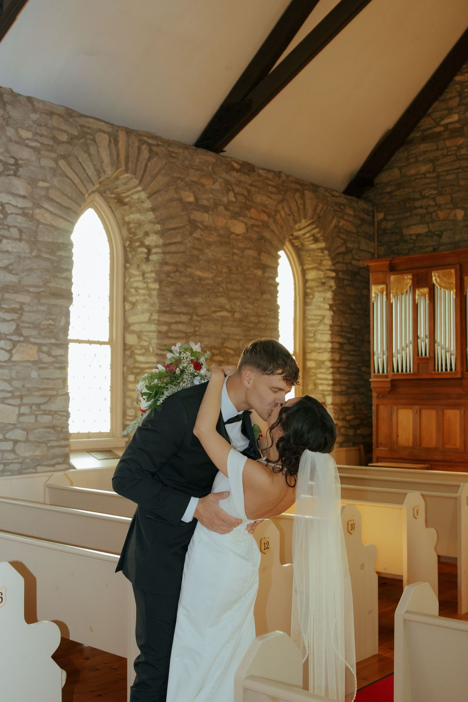 Newlyweds share romantic first kiss near church organ in rustic stone chapel.