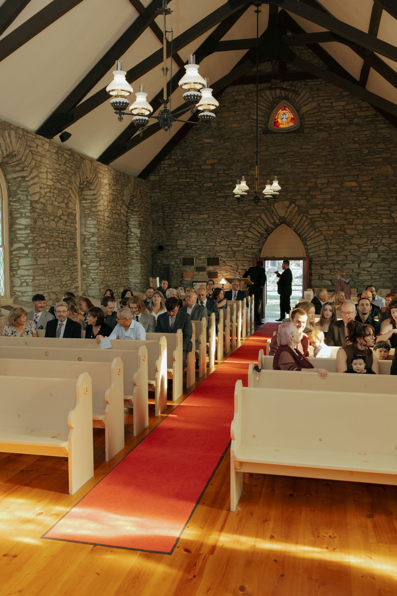 Stone church interior with red carpet aisle, white pews filled with wedding guests, and vaulted ceiling with chandeliers.