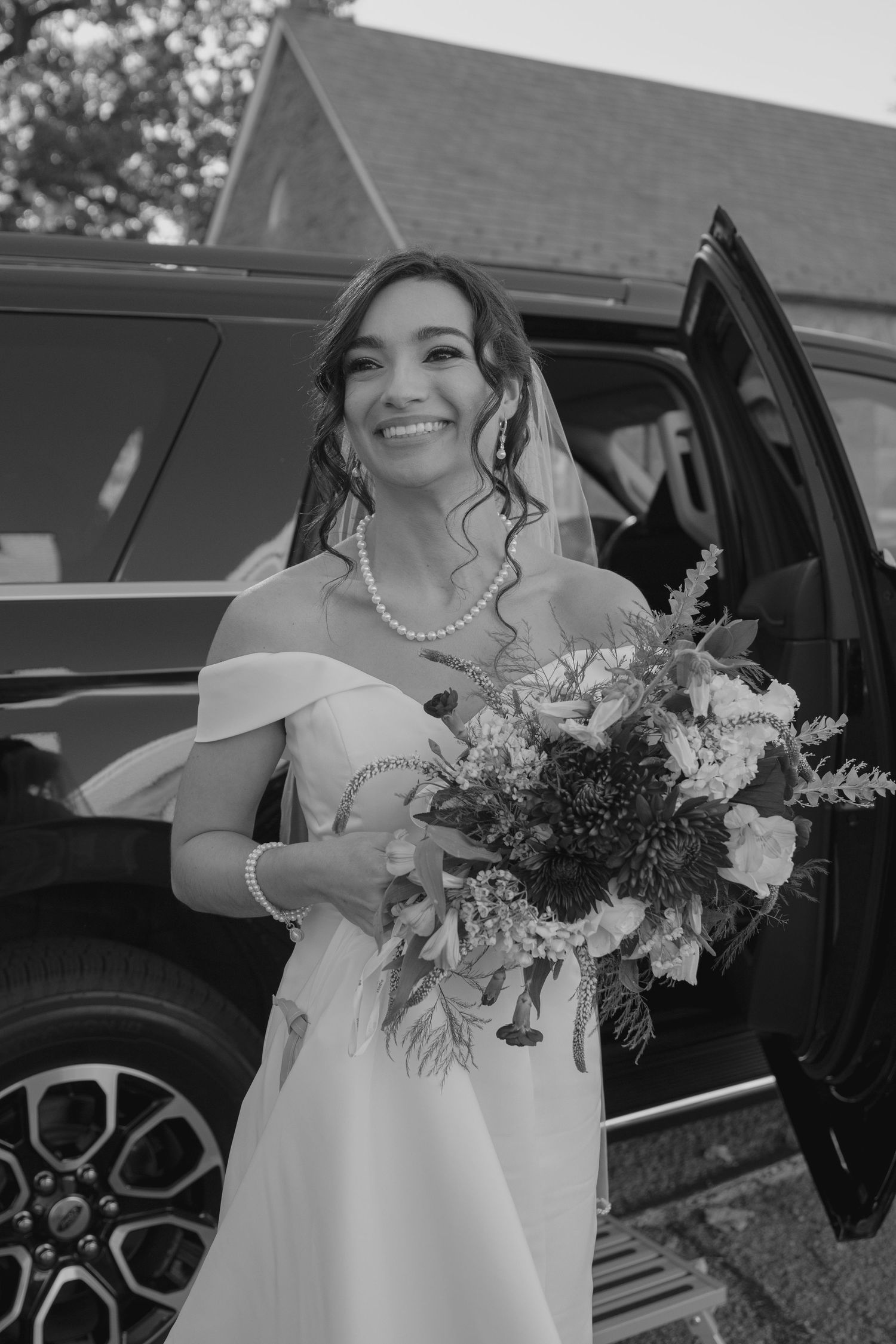 Smiling bride in off-shoulder gown holding bouquet beside luxury SUV on wedding day.