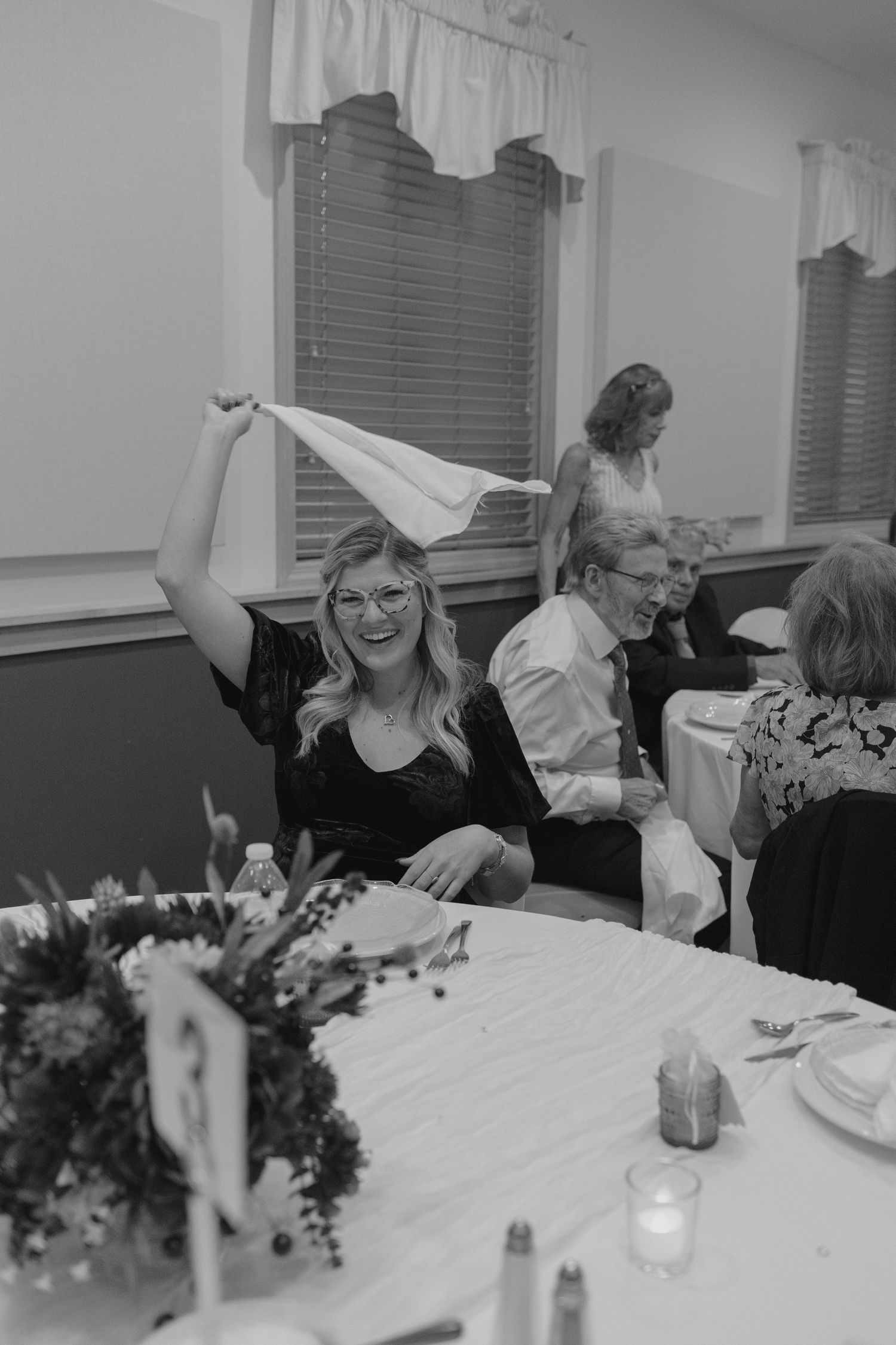 Woman joyfully waves a white napkin at a wedding reception dinner table in black and white photo.