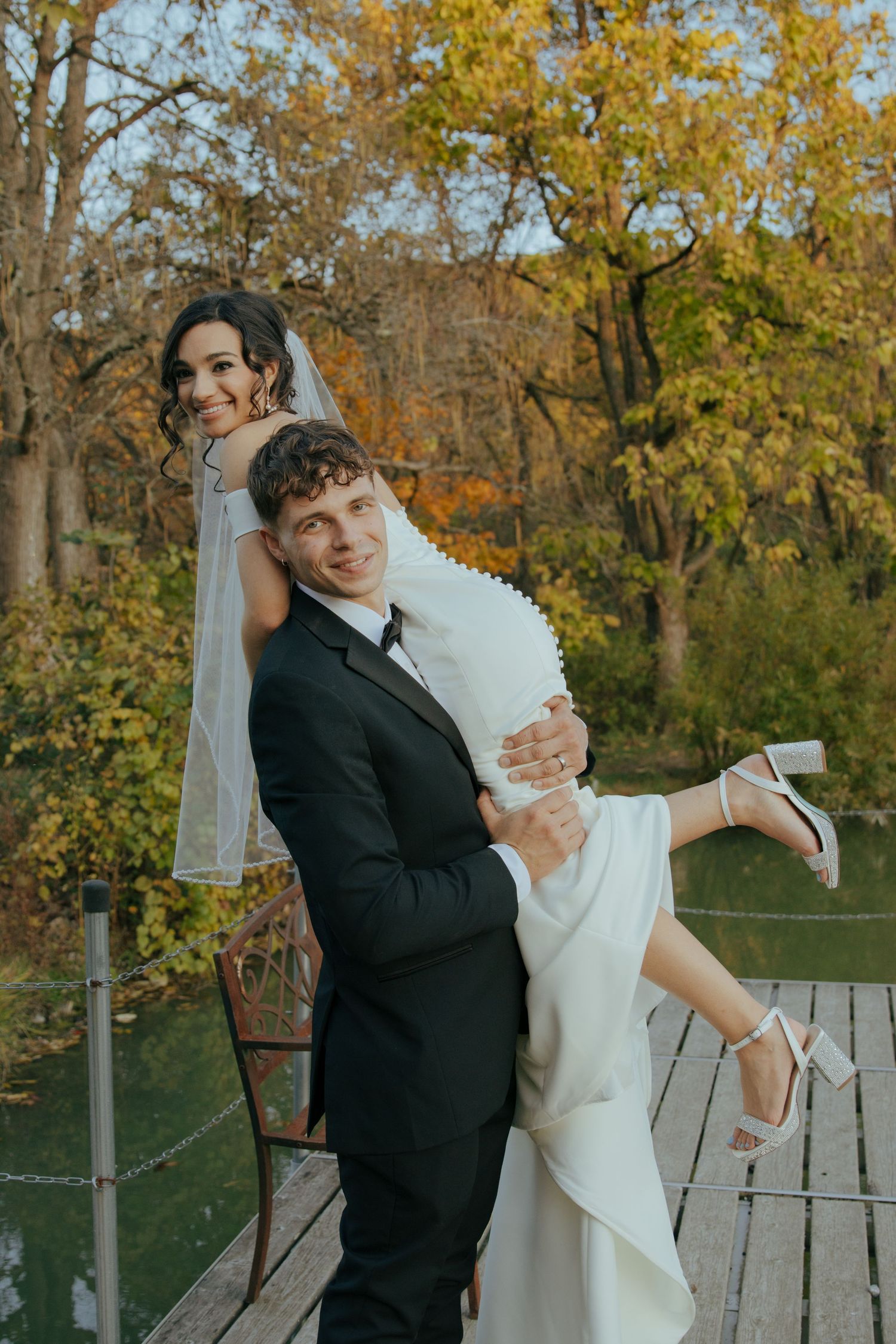 Groom in black suit carries smiling bride in white gown on his back on wooden dock surrounded by autumn trees.