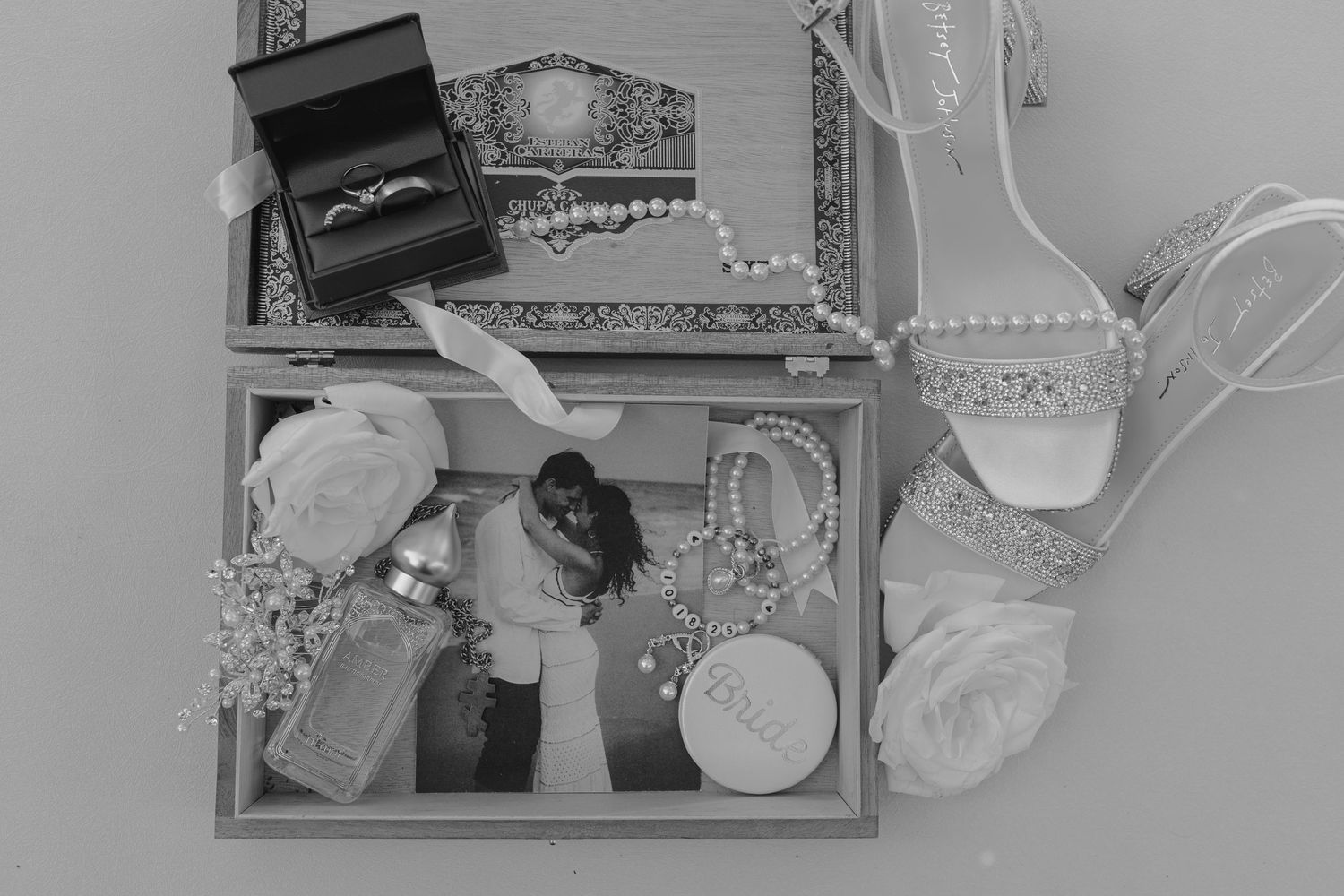 Black and white flat lay of bridal accessories including a ring box, pearl necklace, heels, and a wedding photo.