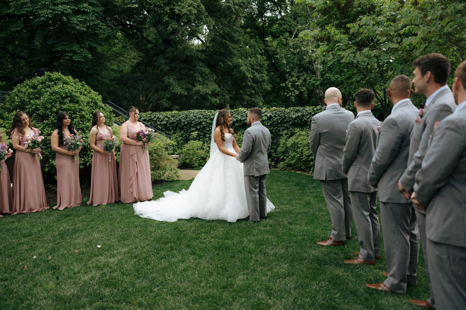 Bride and groom exchange vows outdoors surrounded by bridesmaids in mauve dresses and groomsmen in gray suits.