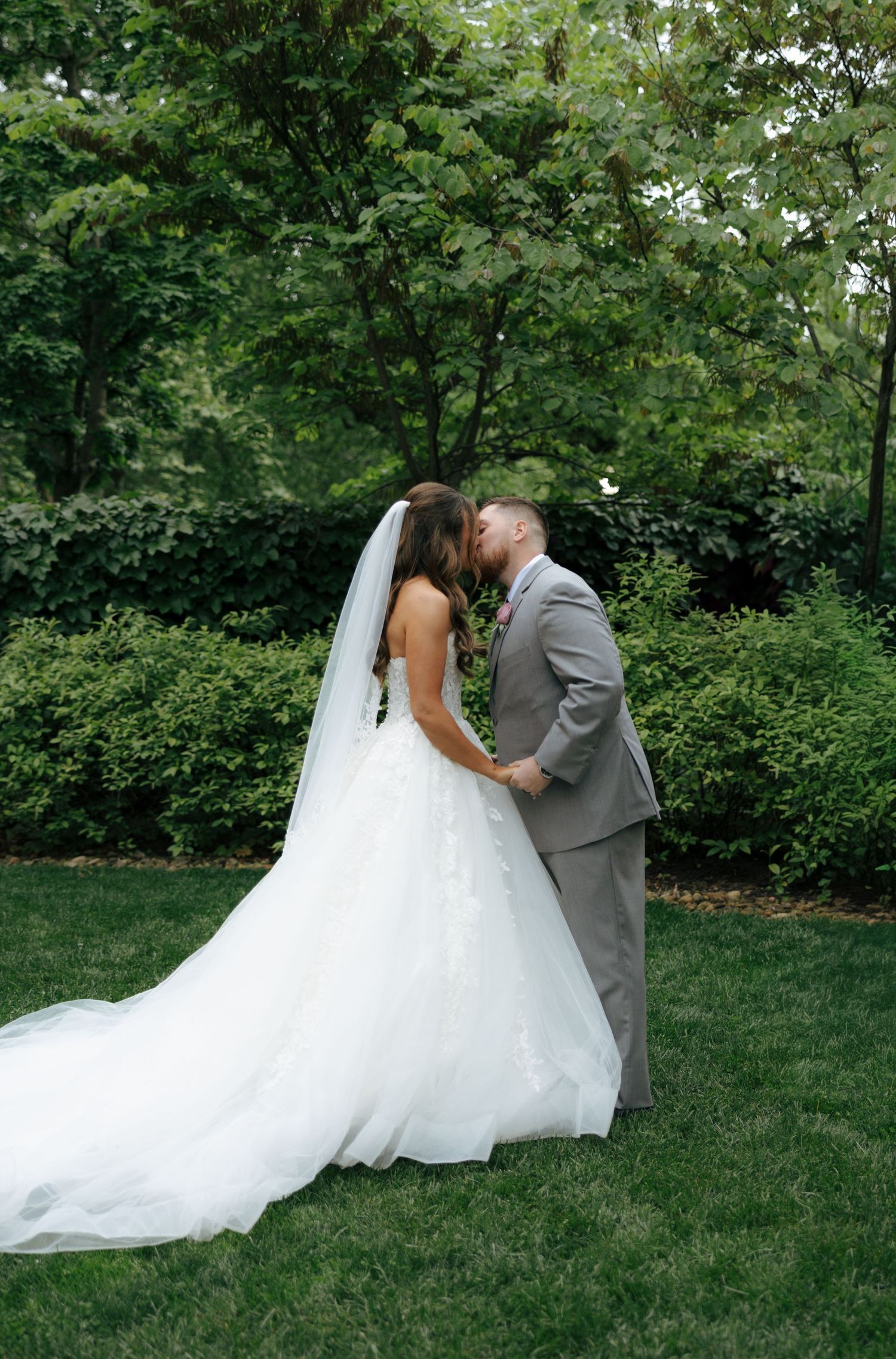 Bride and groom sharing first kiss in garden with long flowing veil and train.
