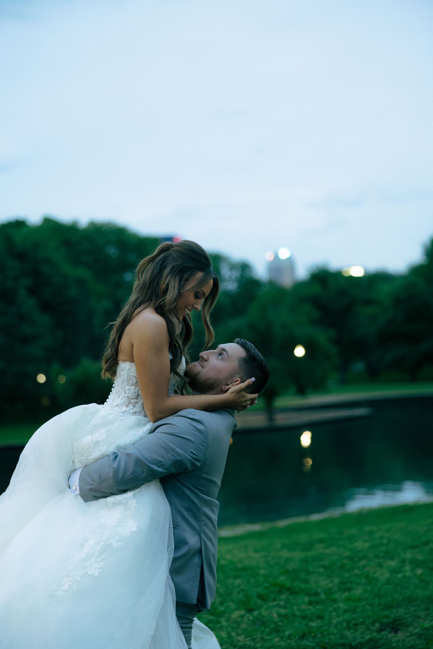 Groom tenderly lifts smiling bride in lace wedding gown near reflective pond at twilight.
