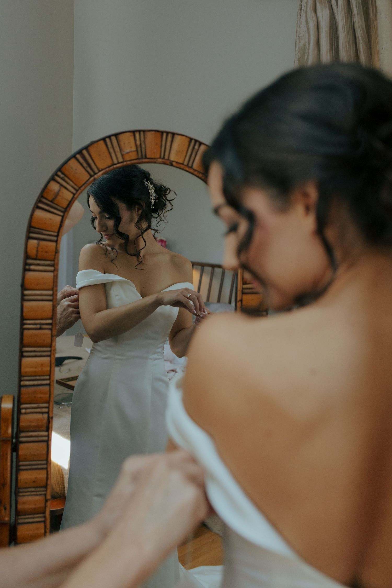 Bride in off-shoulder white gown adjusting dress while viewing reflection in ornate mirror.
