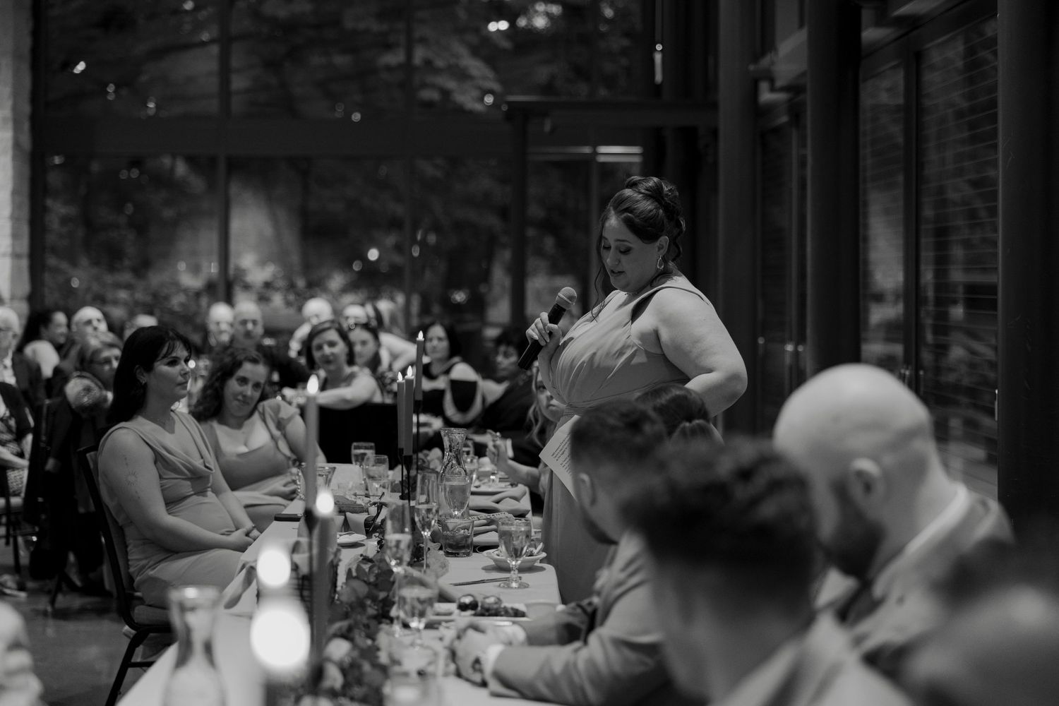 Woman giving a speech at an elegant wedding reception dinner in a candlelit venue with guests seated at long tables.