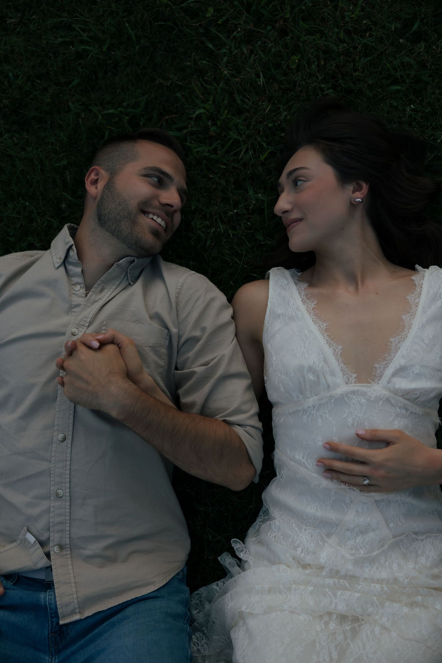 Couple lying on grass holding hands and smiling at each other, woman in white lace dress with engagement ring.