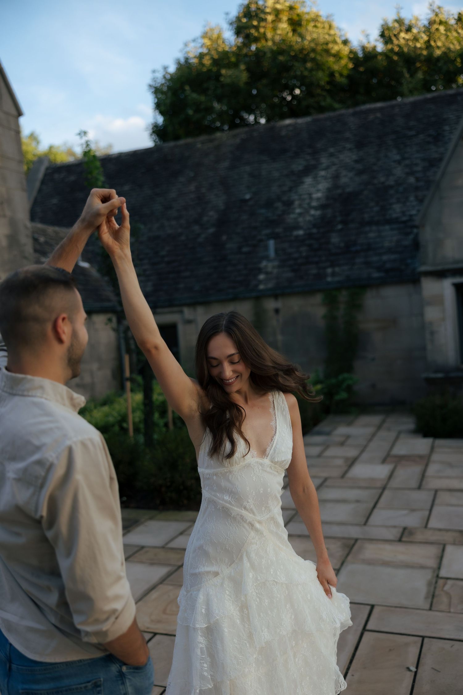 Bride in white lace dress twirls as groom spins her on stone patio of historic venue.