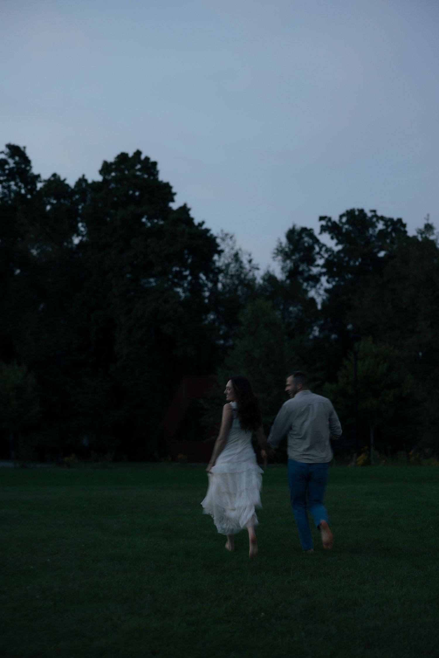 Couple holding hands running across a dark green lawn at twilight with trees in background.