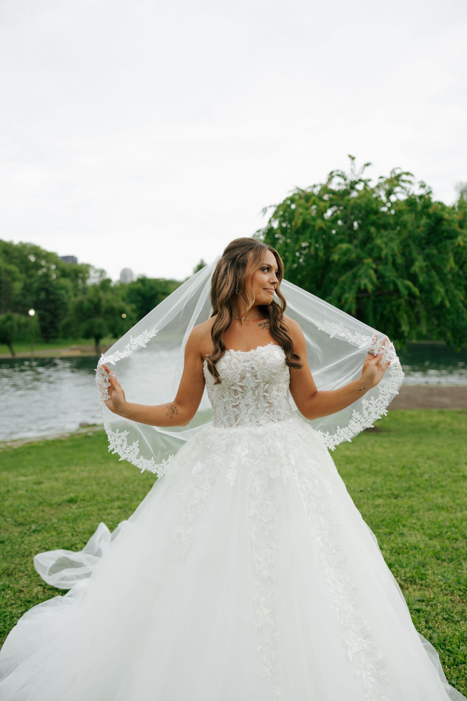 Elegant bride in a lace ball gown lifting her veil near a serene lake on her wedding day.
