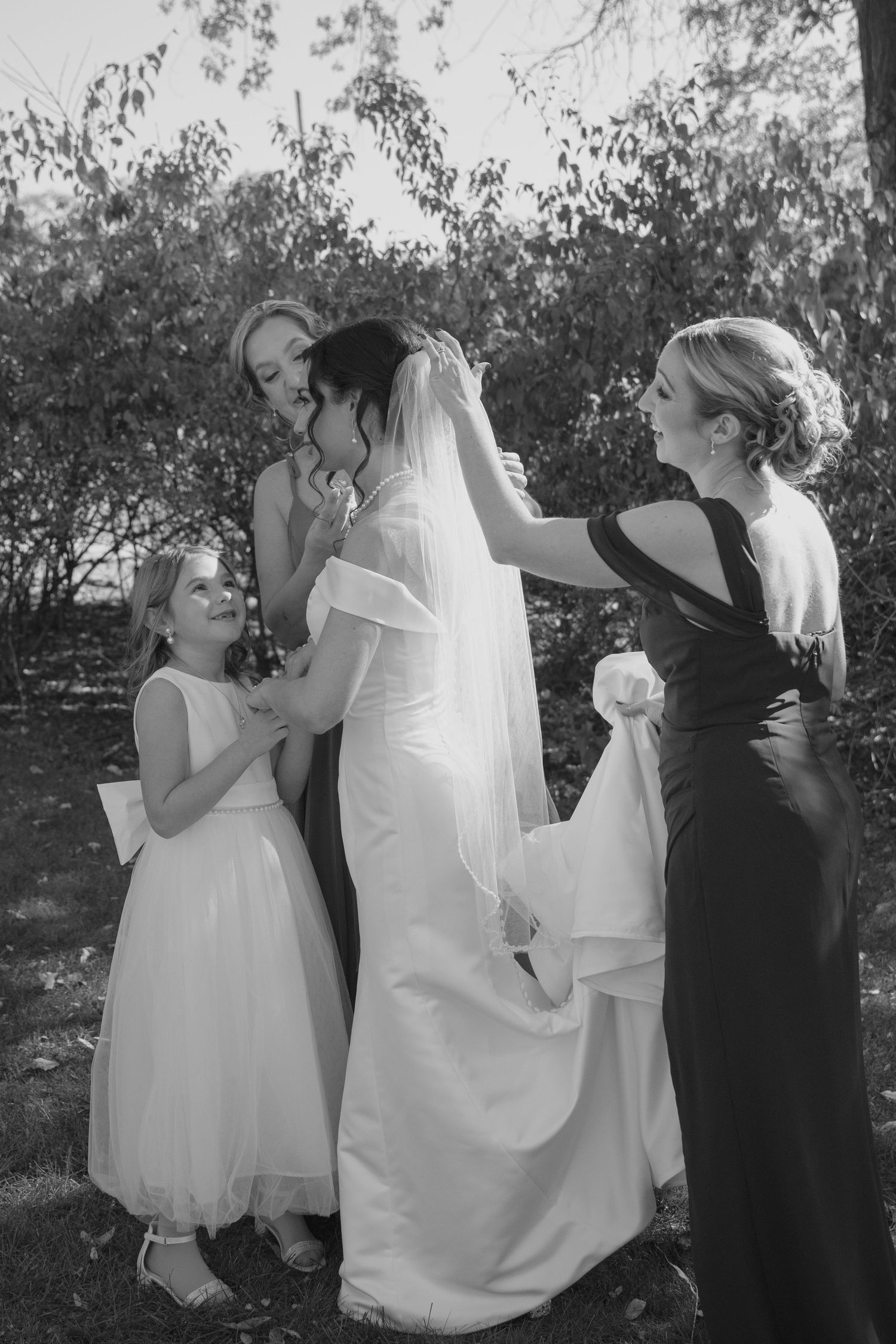 Black and white photo of bridesmaids and flower girl adjusting the brides veil outdoors before the wedding.
