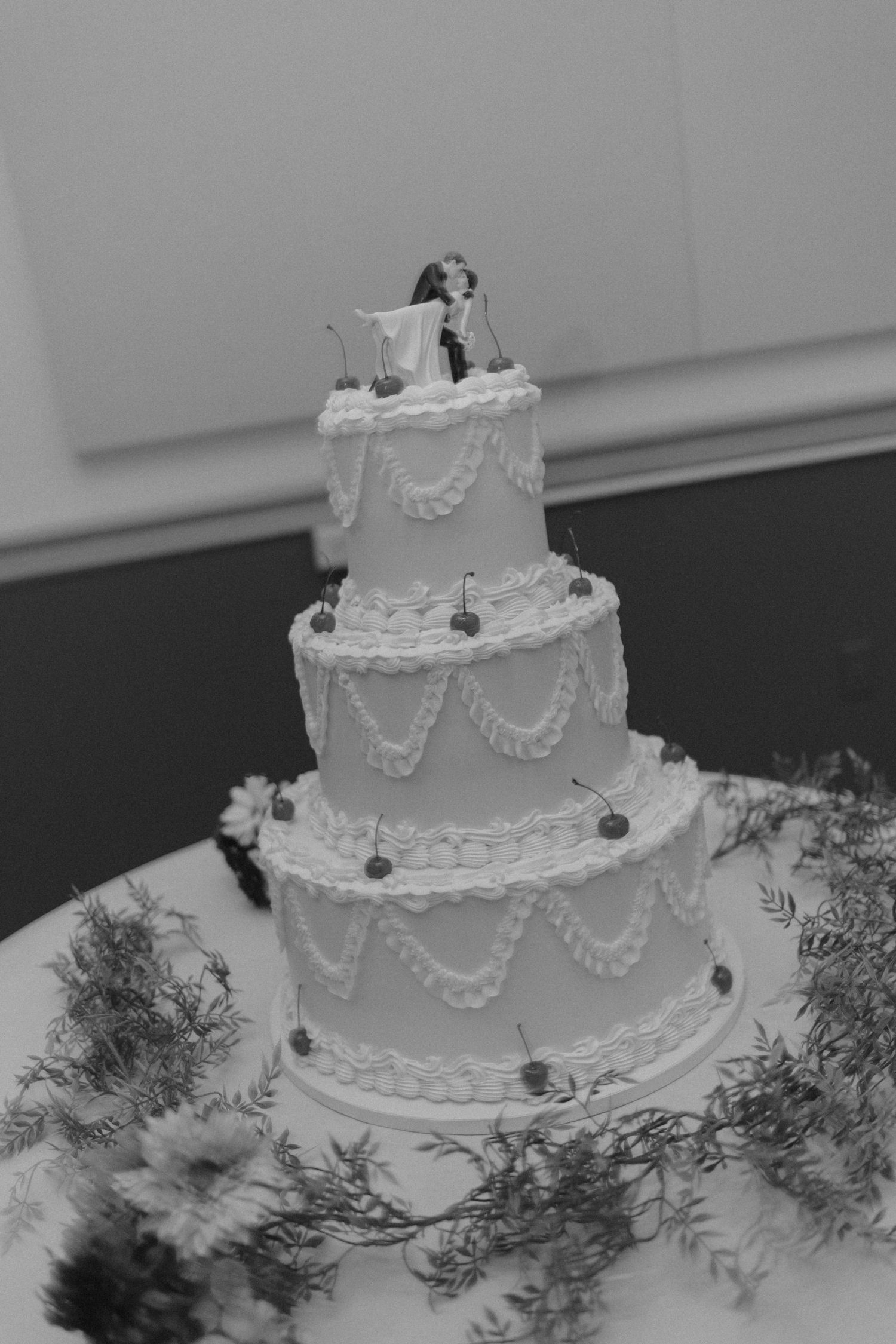 Elegant three-tier white wedding cake with bride and groom topper and floral decorations in black and white.