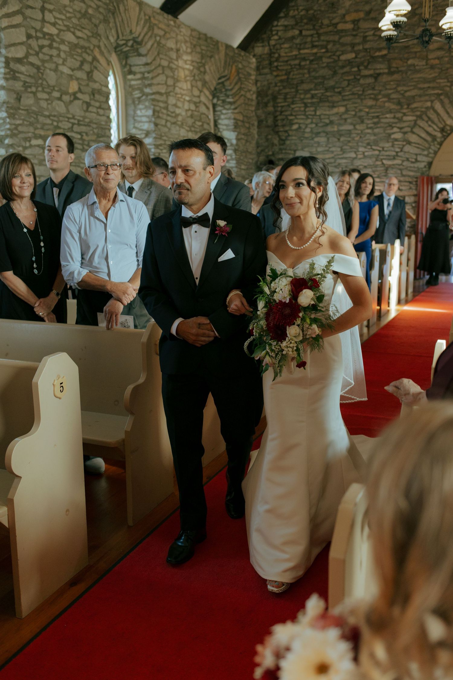 Bride and groom walk down red carpet aisle in stone church, bride holding white and red bouquet.