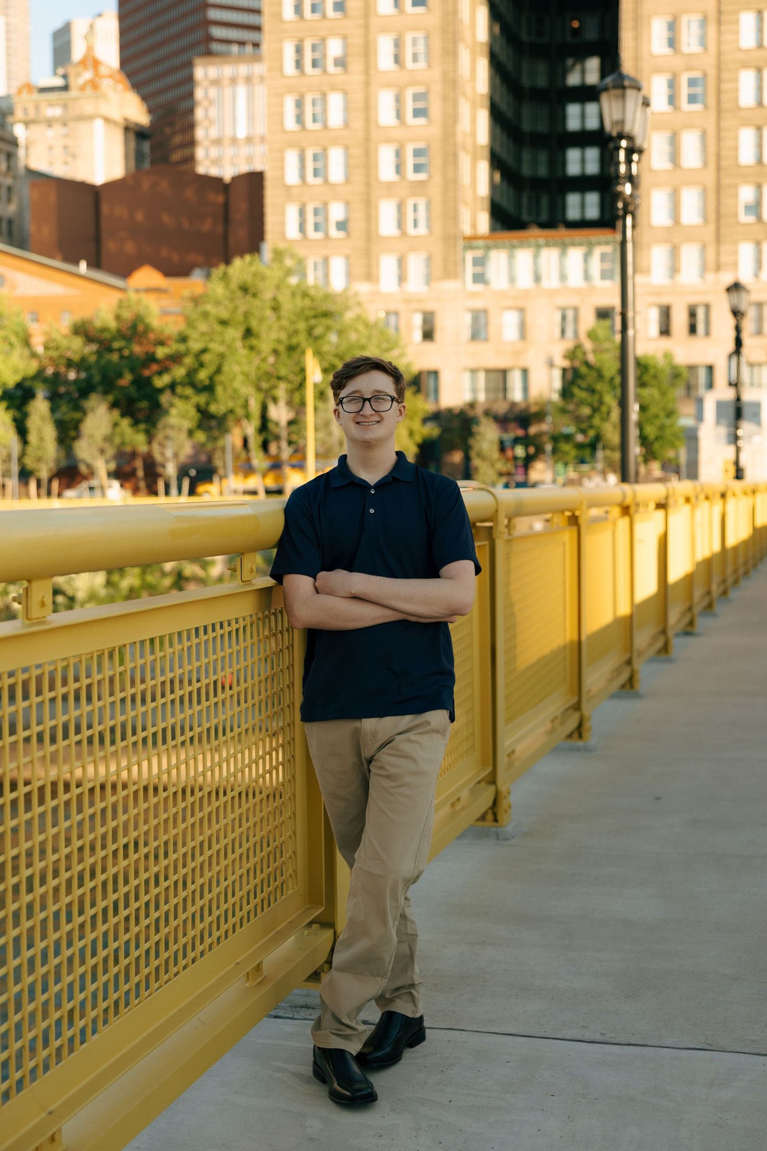 Young man in navy polo with arms crossed leaning against yellow bridge railing, urban backdrop.