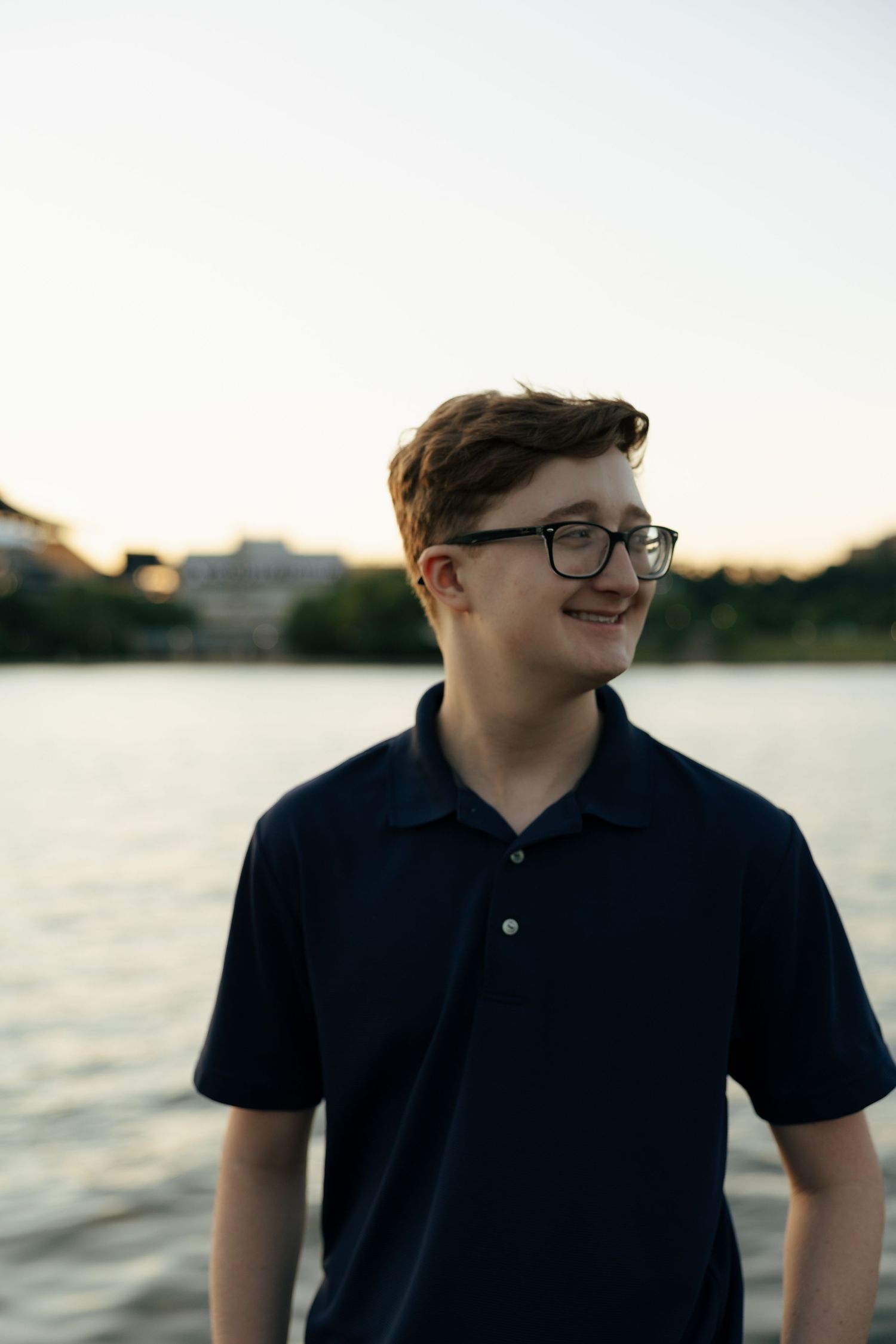 Young man in dark polo shirt and glasses smiling by a lake at sunset.