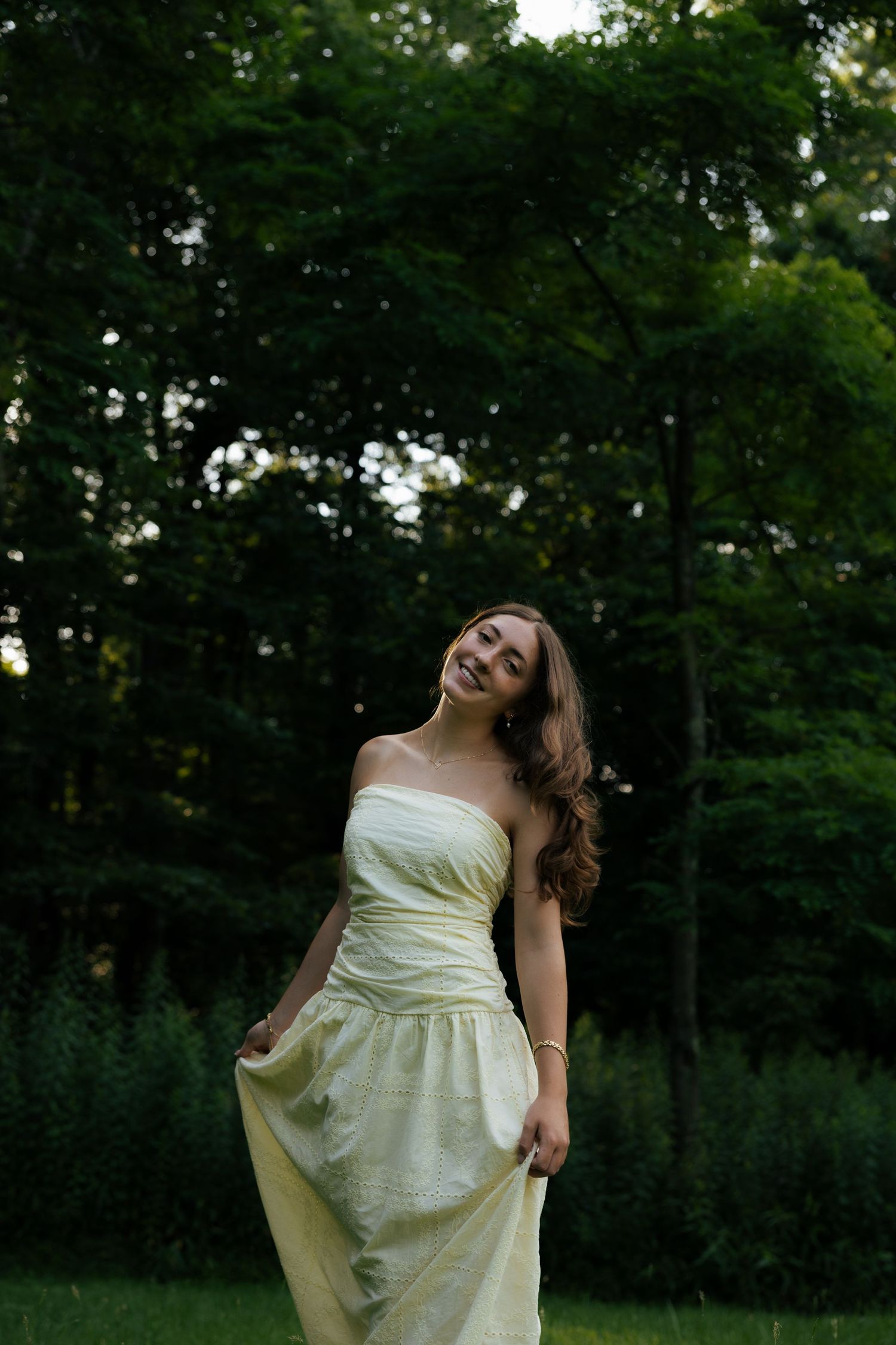 Young woman in strapless yellow gown smiling outdoors in lush green garden setting.