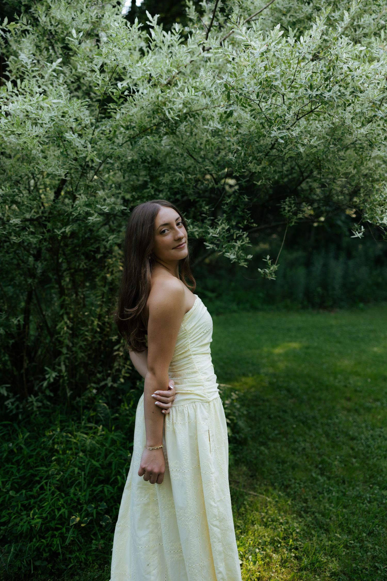 Young woman in a flowing yellow strapless dress standing near white flowering bushes in a garden.