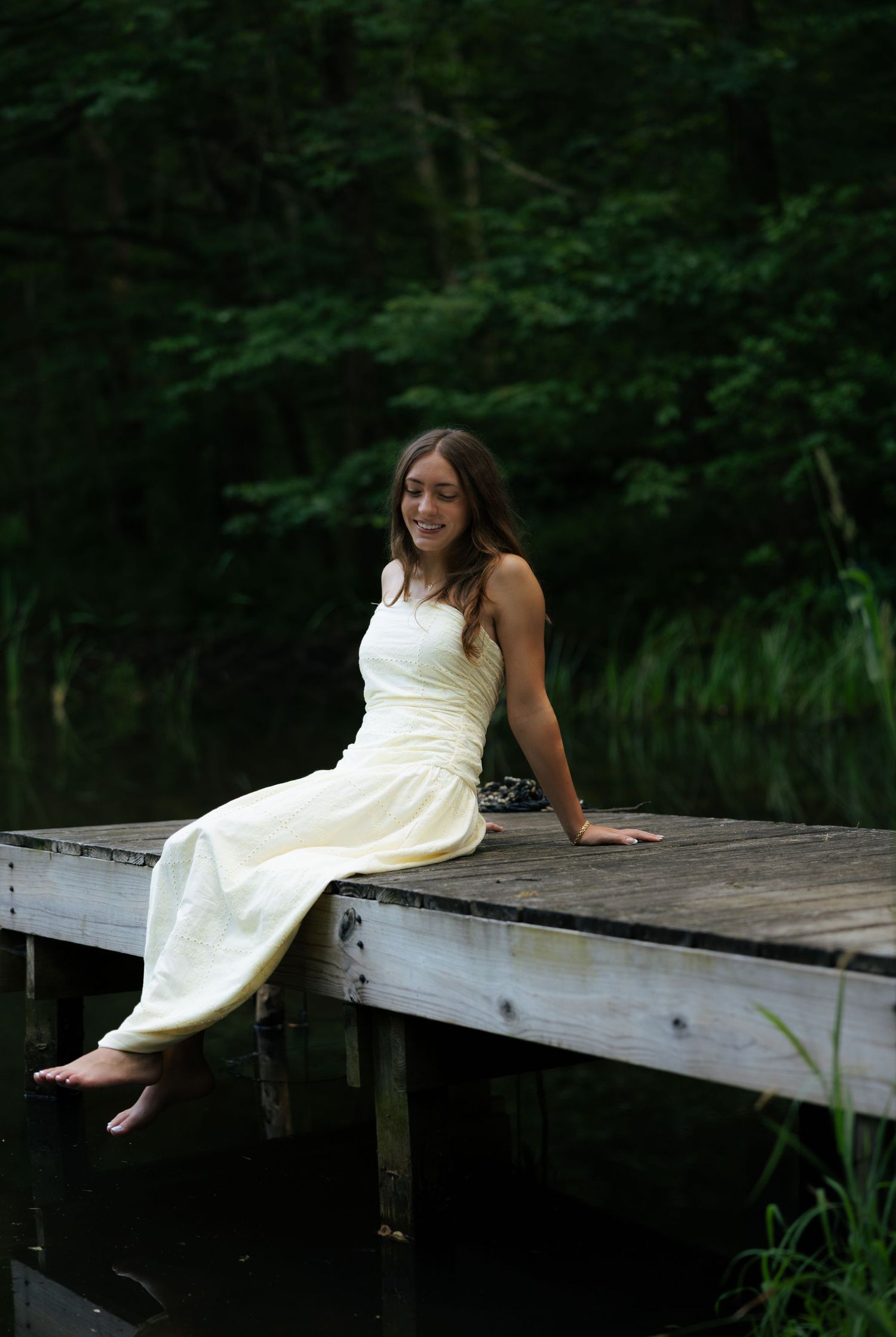 Young woman in yellow dress sitting barefoot on wooden dock over calm lake in forest.